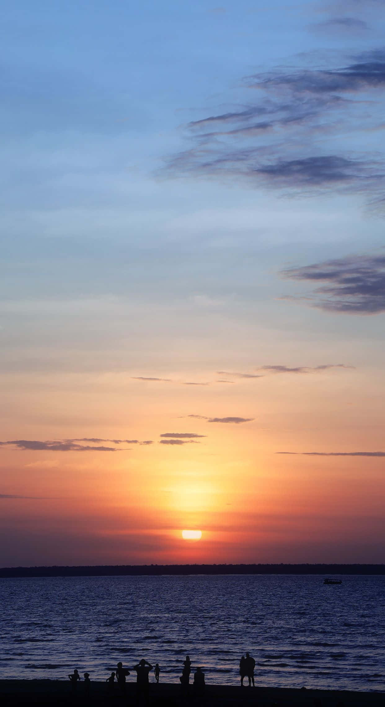 Coastal Sunset 桌布 壁紙 — 剪影人物觀看鮮豔的橙色太陽在平靜的海面下沉，天空是柔和的藍色，散佈著雲朵。