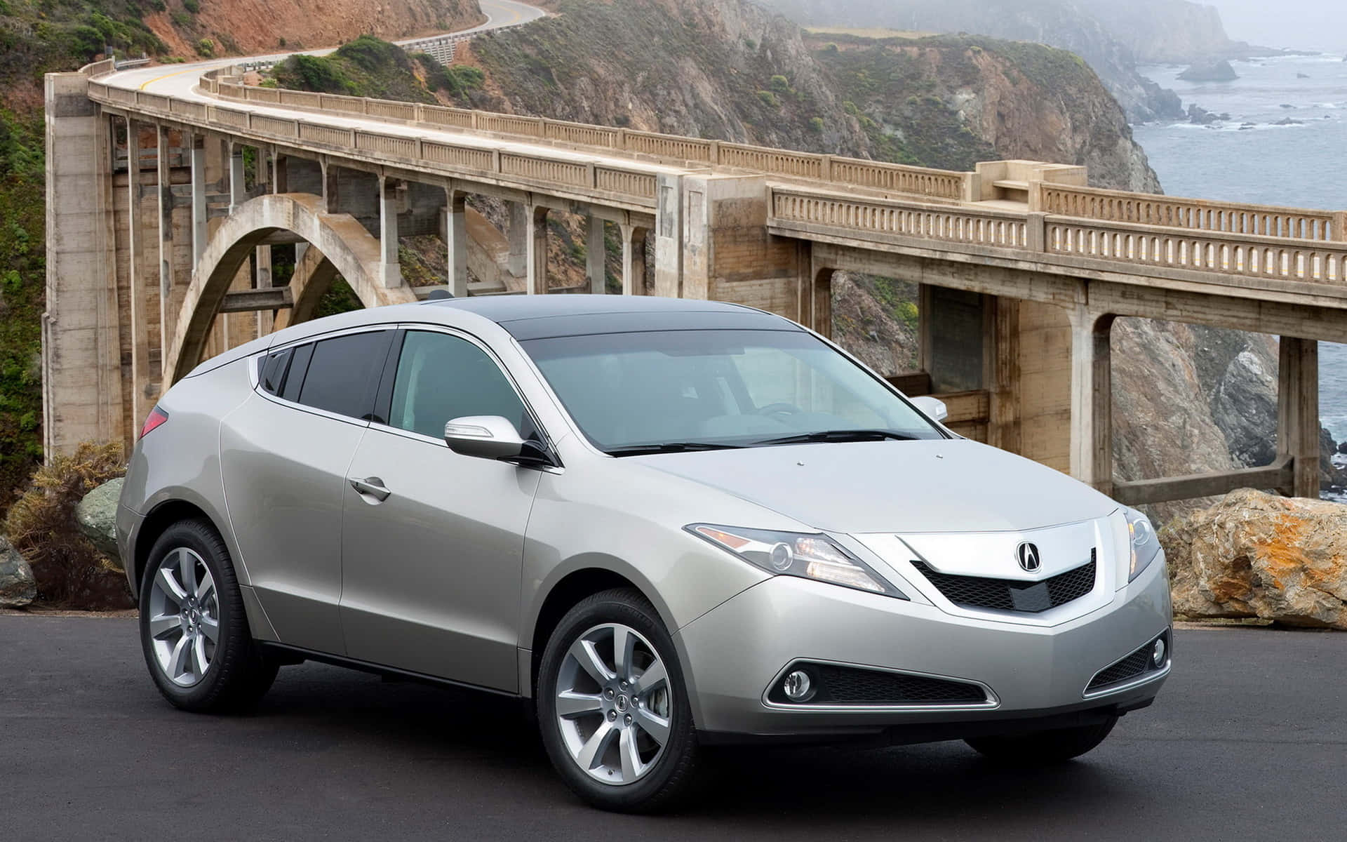 Acura Zdx 桌布 壁紙 — 一輛銀色Acura SUV停泊在海岸公路上，位於石拱橋下方，背景有海洋懸崖和波浪。