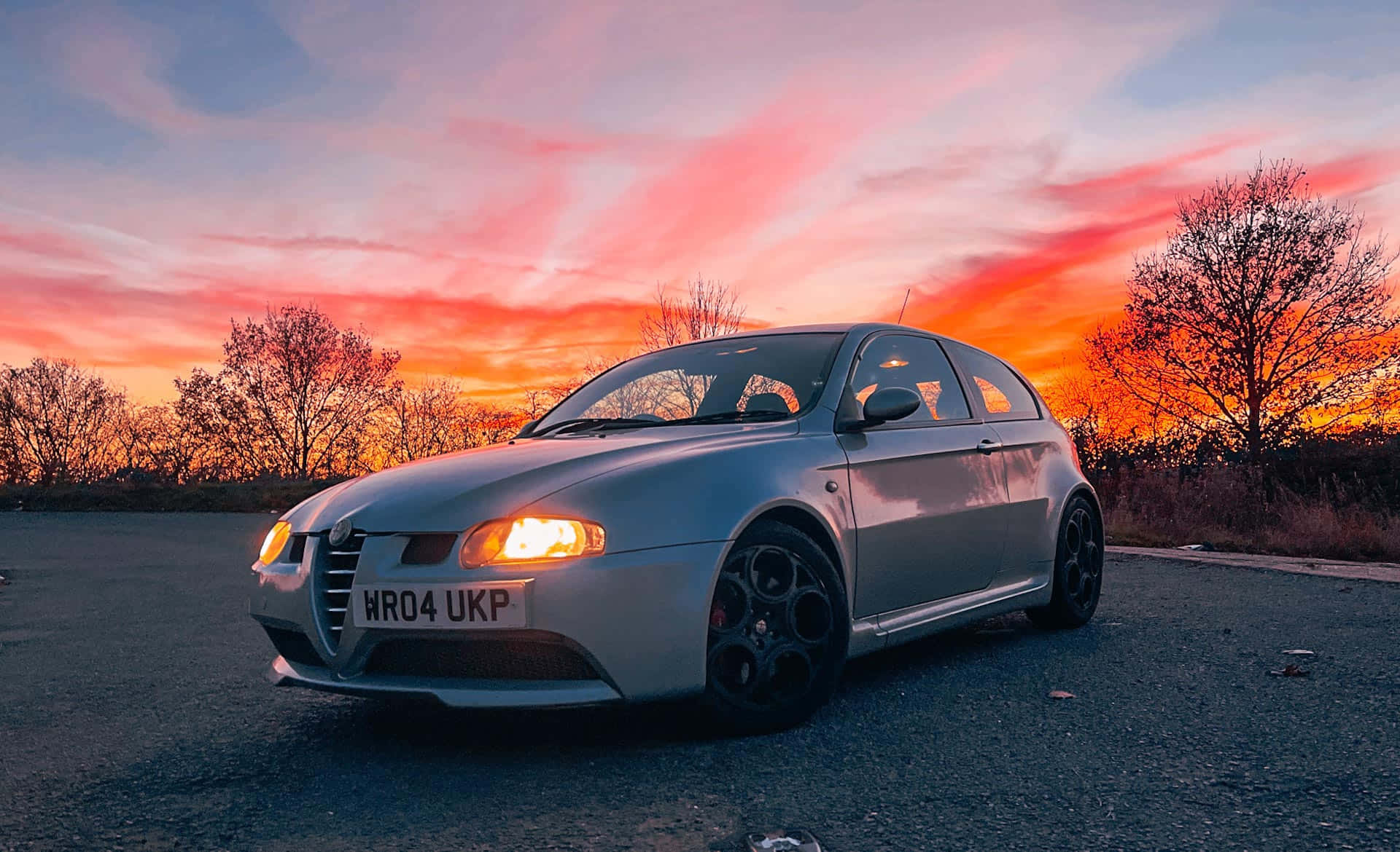 Alfa Romeo 147 桌布 壁紙 — 一輛銀色愛快羅密歐跑車停泊在瀝青路上，背景是火紅的日落天空，襯托著光禿樹木的剪影。