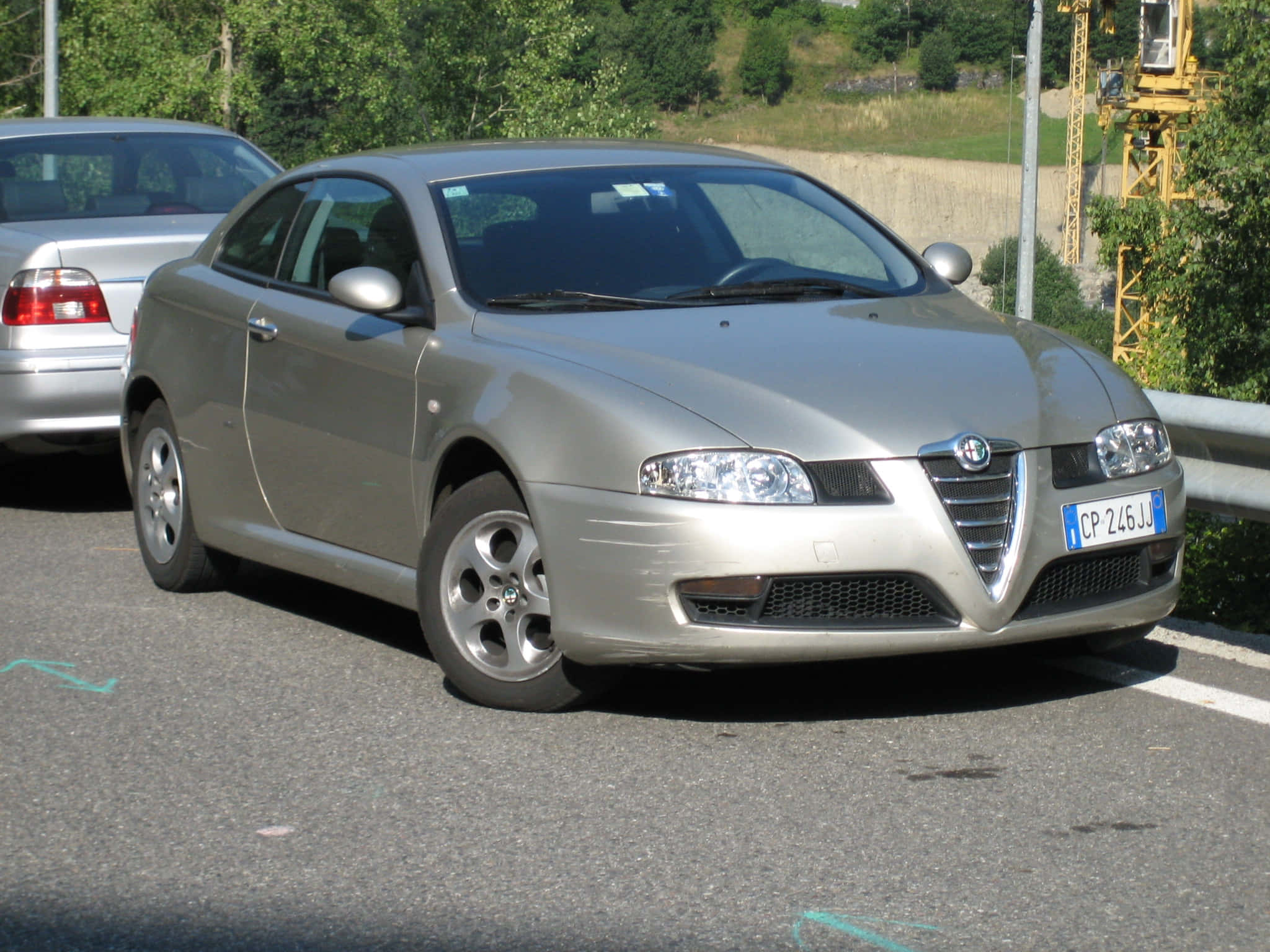 Alfa Romeo Gt 桌布 壁紙 — 一輛銀色愛快羅密歐Spider雙門跑車停泊在陽光普照的路邊，背景有綠樹和黃色起重機。