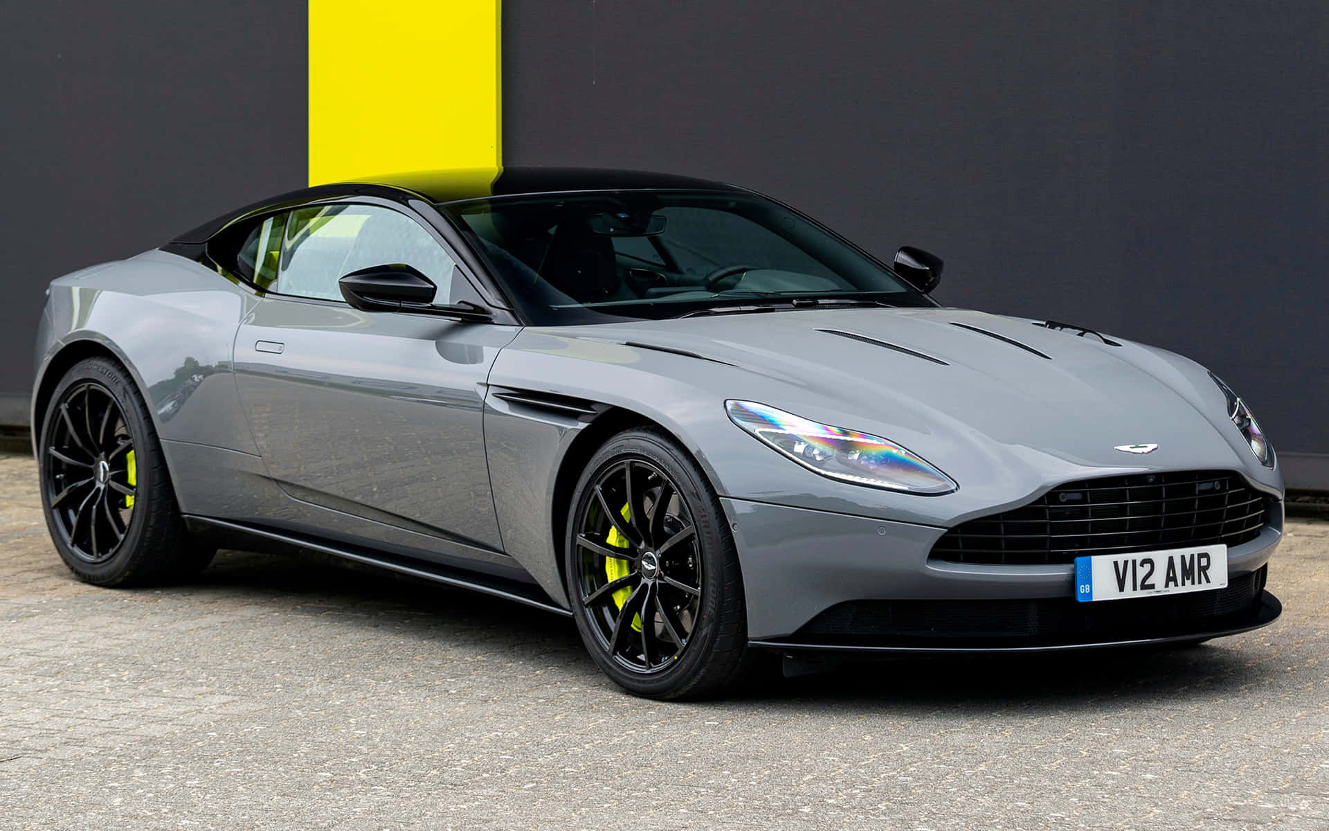 Aston Martin Db11 桌布 壁紙 — 一輛時尚的銀色 Aston Martin DB11 AMR，帶有黑色飾邊和鮮黃色制動卡鉗，停放在大膽的黃黑牆前。