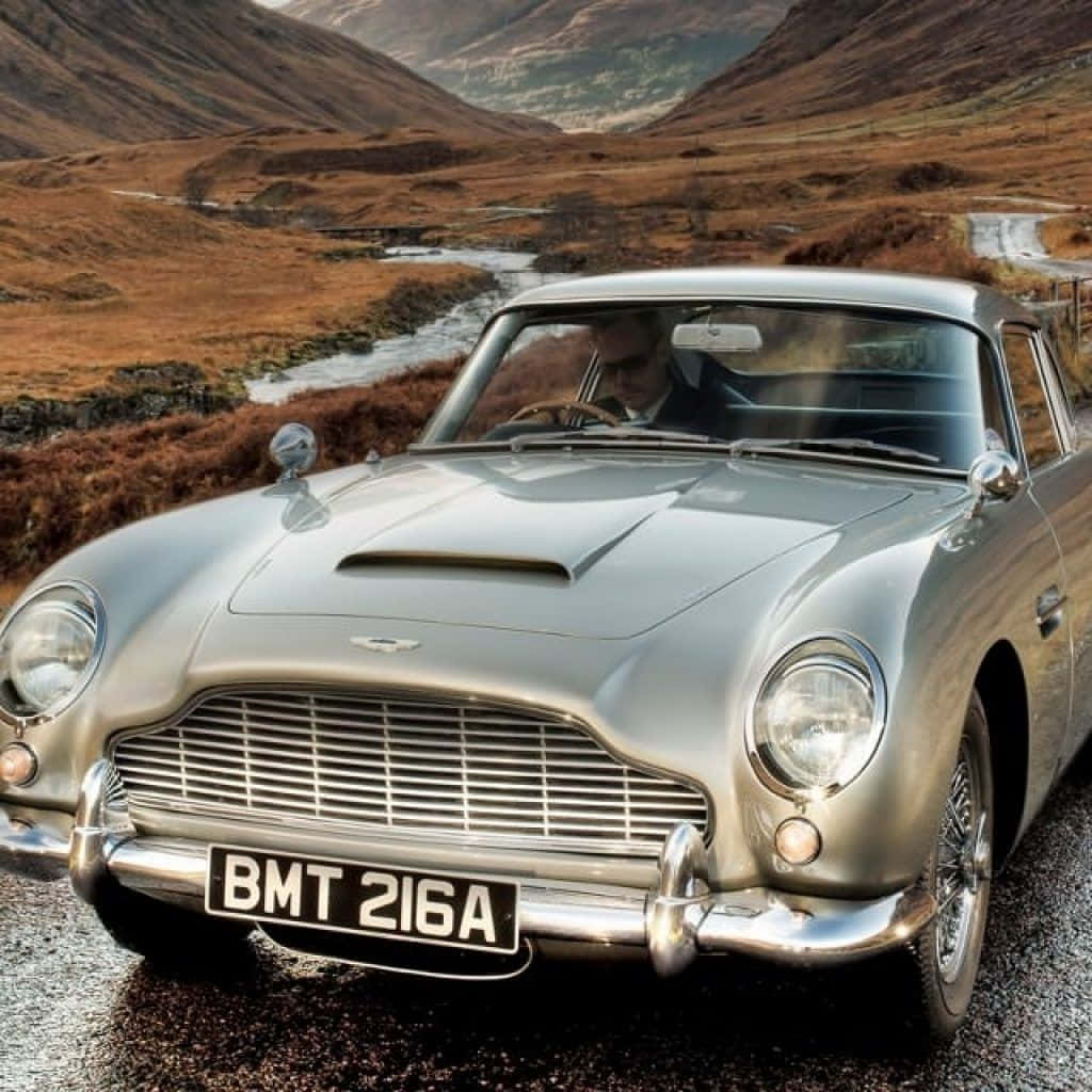 Aston Martin Db5 桌布 壁紙 — 一輛銀色Aston Martin DB5在濕滑的山路上閃閃發光，河流蜿蜒穿過金棕色的山丘。
