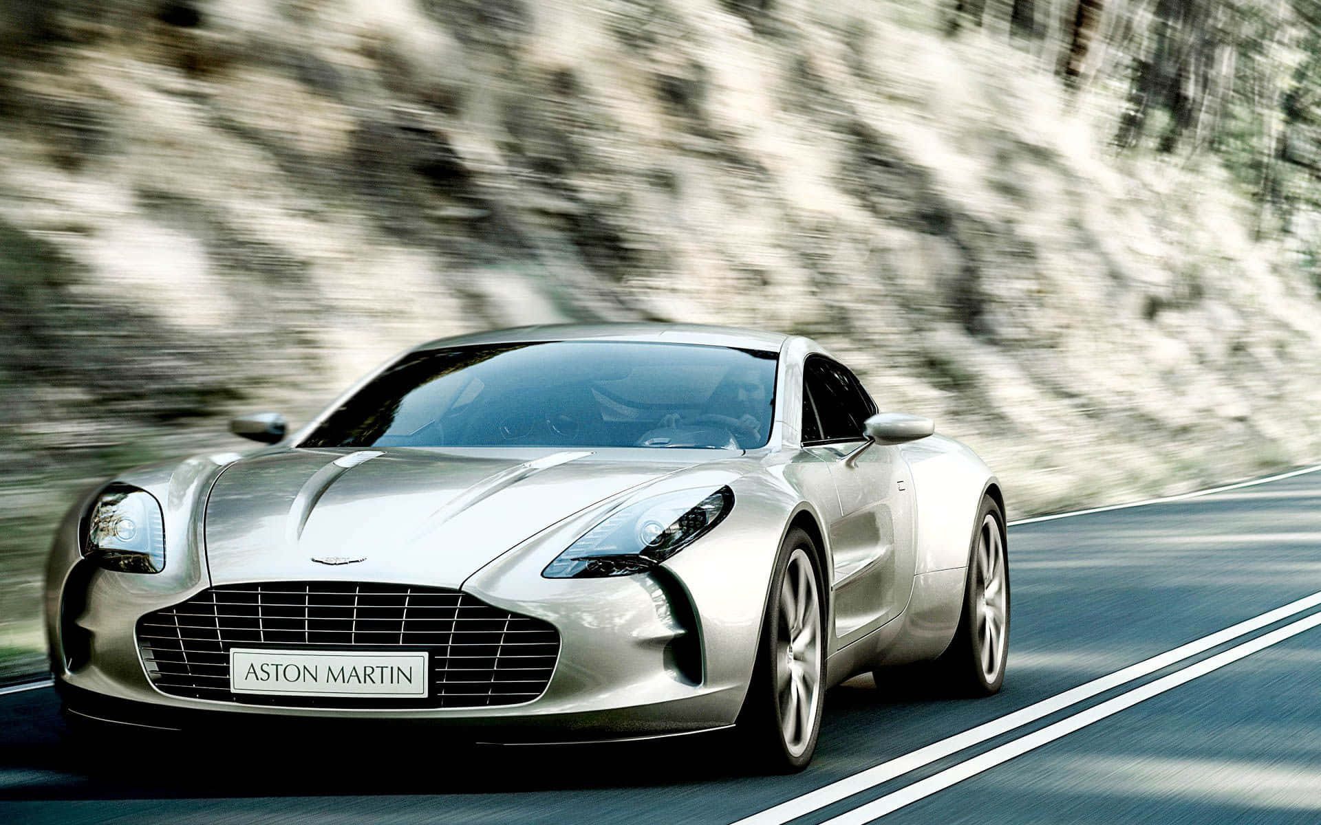 Aston Martin One-77 桌布 壁紙 — 一輛銀色Aston Martin跑車在蜿蜒的山路上疾馳，背景有模糊的樹葉。