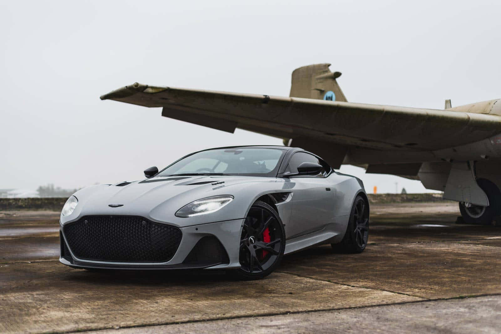 Aston Martin Dbs Superleggera 桌布 壁紙 — 一架時尚的銀色Aston Martin跑車停泊在濕滑的停機坪上，旁邊是一架大型復古軍用飛機，天空多雲。