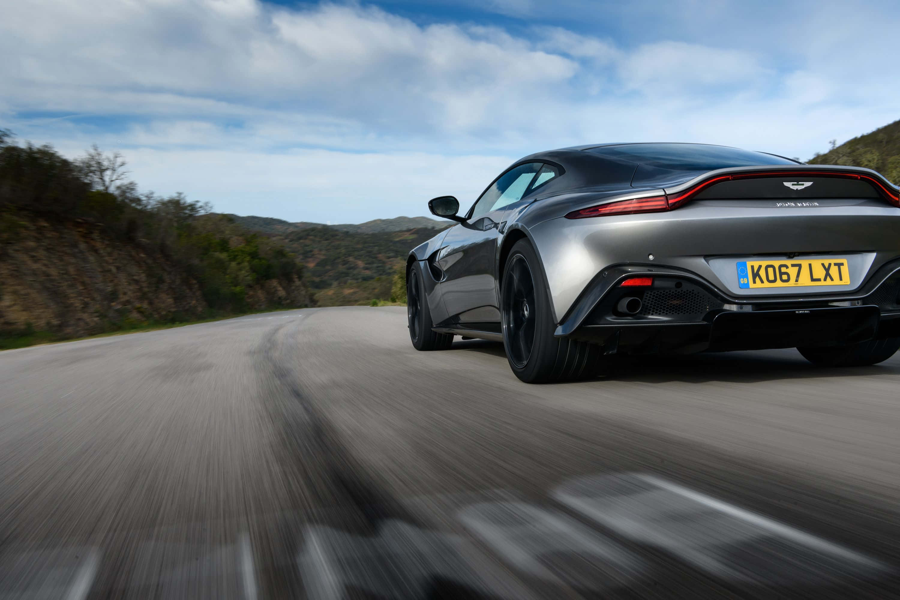 Aston Martin Vantage 桌布 壁紙 — 一輛流線型銀色Aston Martin Vantage在部分多雲的天空下沿著蜿蜒的山路疾馳。