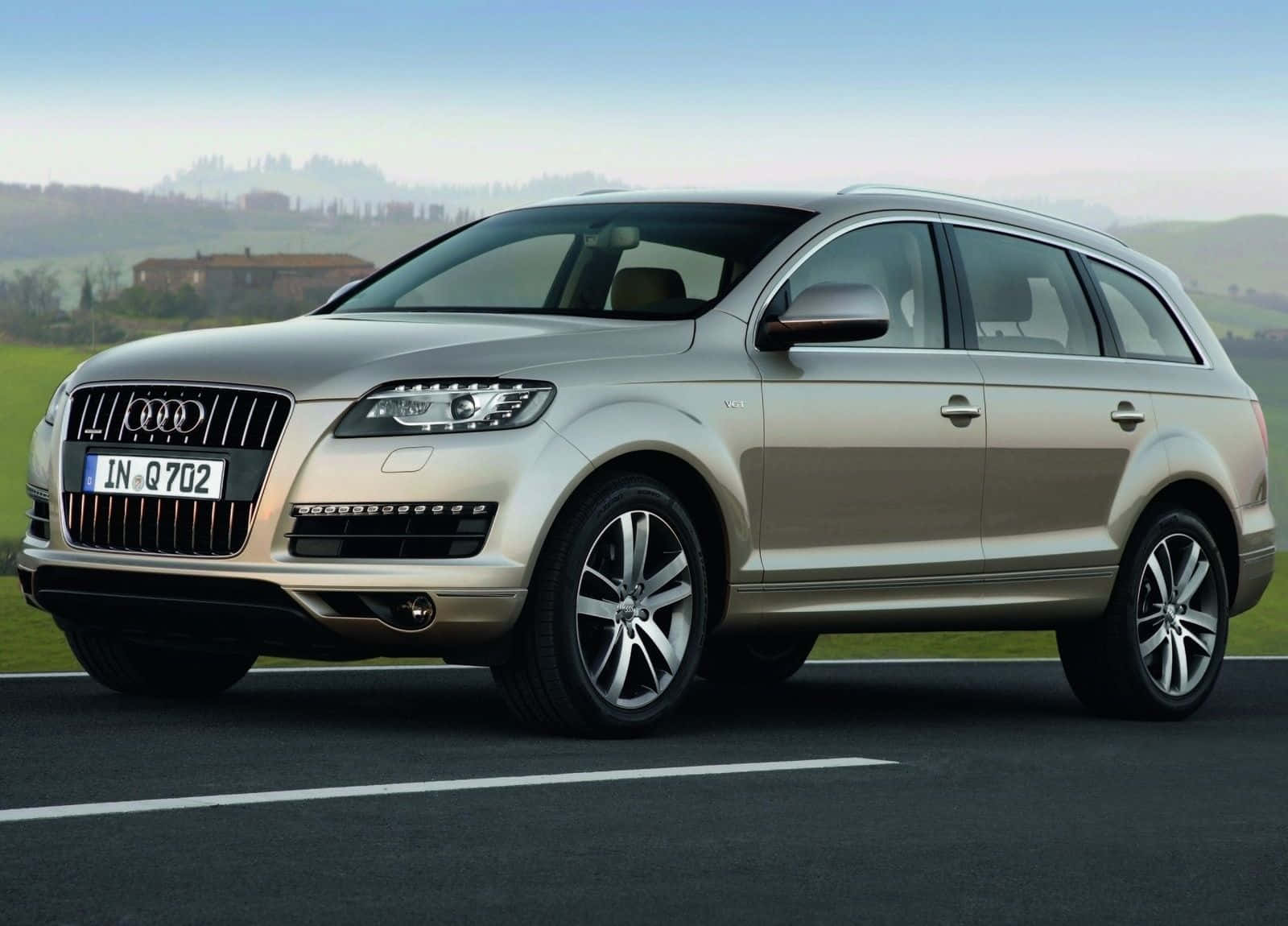 Audi Q7 桌布 壁紙 — 一輛時尚的銀色奧迪Q7 SUV停泊在瀝青路上，周圍是連綿起伏的綠色山丘，遠處有一座農舍，天空清澈湛藍。