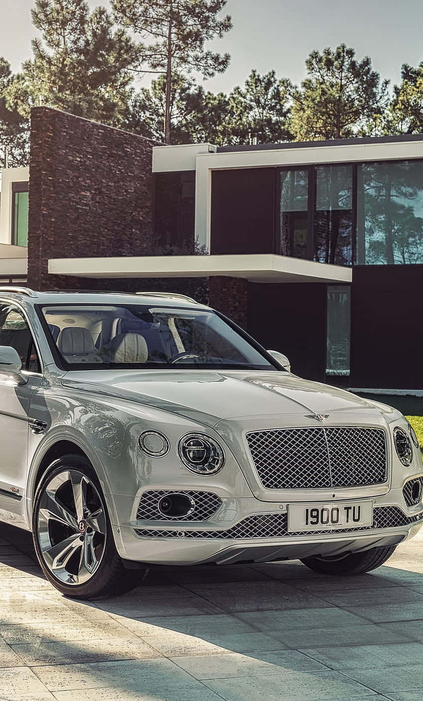 Bentley Bentayga 桌布 壁紙 — 一輛時尚的銀色賓利Bentayga SUV停泊在鋪有瓷磚的車道上，前面是一座現代化的石頭和玻璃建築房屋，周圍環繞著高大的松樹。