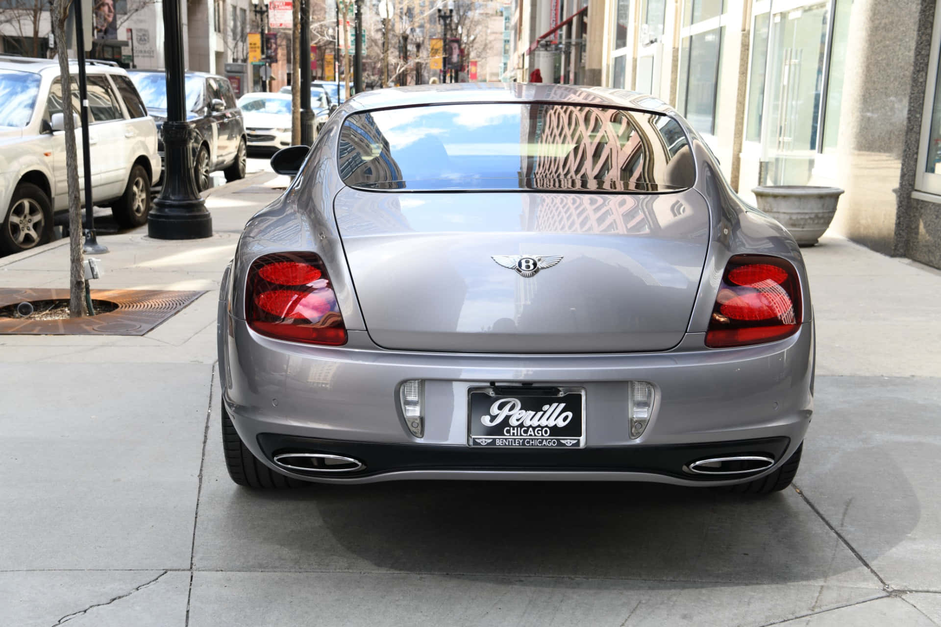 Bentley Continental Supersports 桌布 壁紙 — 一架銀色賓利 Continental GT 停泊在城市行人路上，其光亮的後窗反射出裝飾圍欄和城市建築。