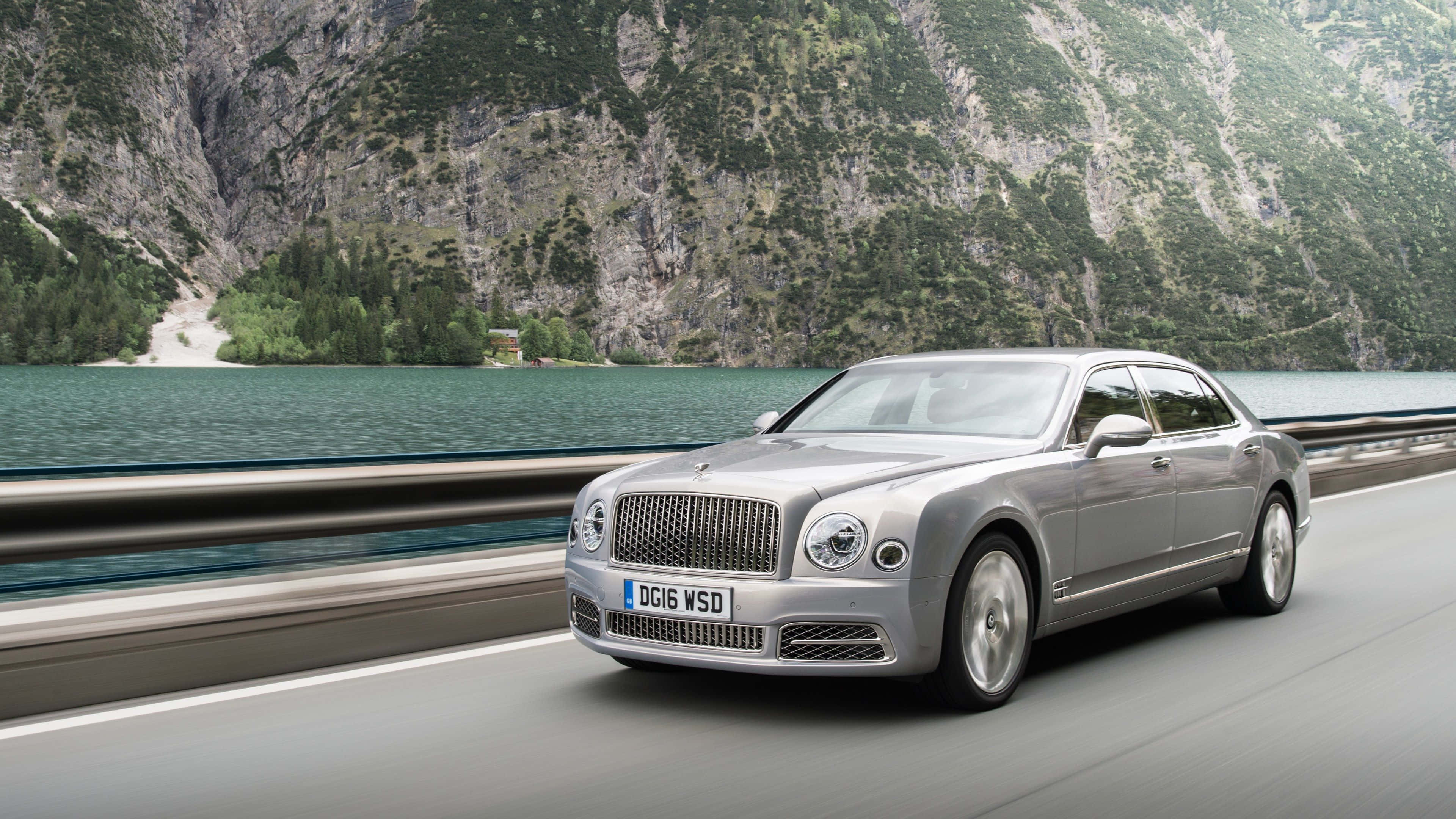 Bentley Mulsanne 桌布 壁紙 — 一輛銀色賓利慕尚沿著山路行駛，旁邊是寧靜的碧綠湖泊，背景是森林覆蓋的懸崖。