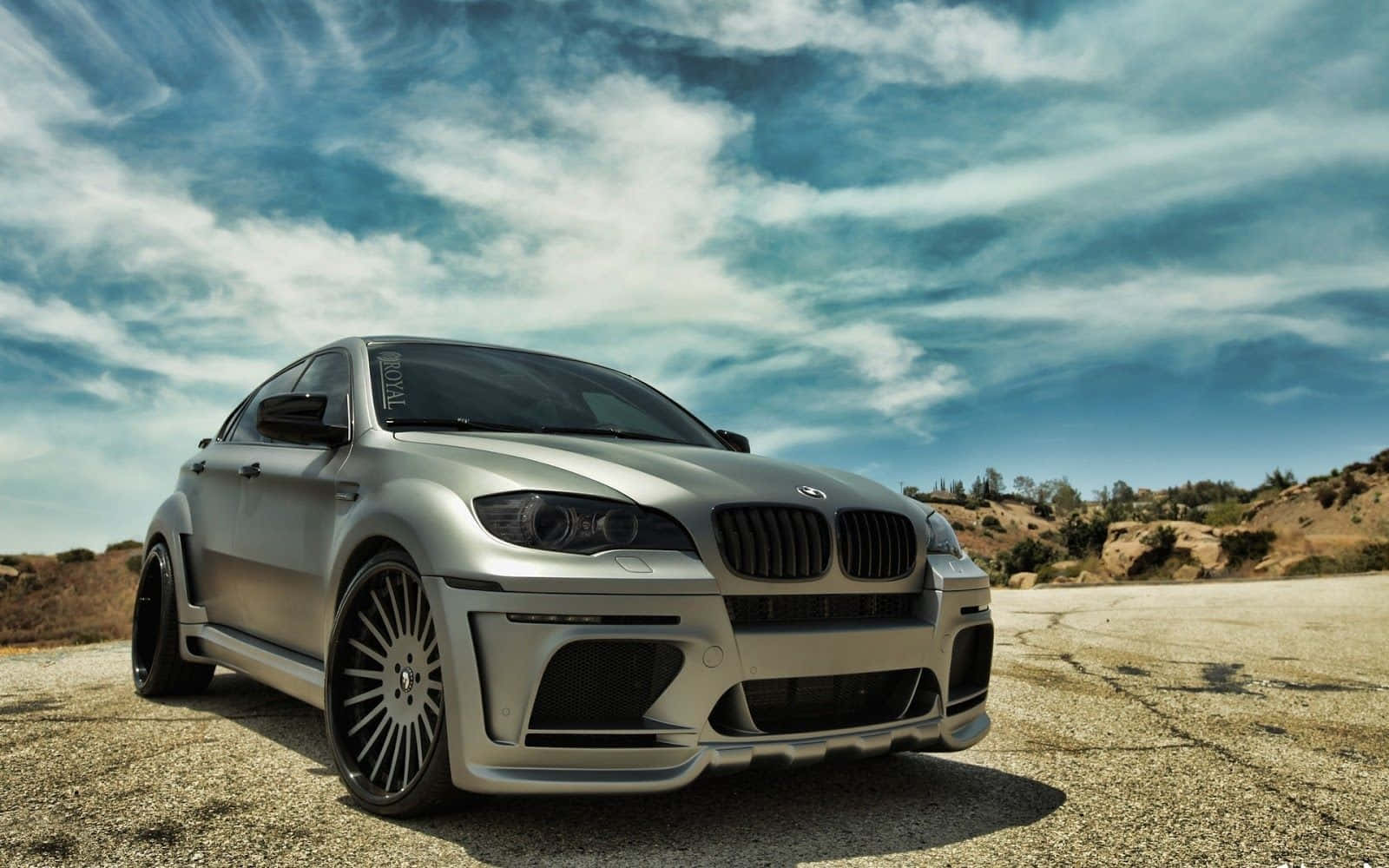 Bmw X6 桌布 壁紙 — 一輛銀色BMW M6雙門跑車配備定制寬體套件，停在裂開的瀝青路面上，天空多雲而戲劇化。