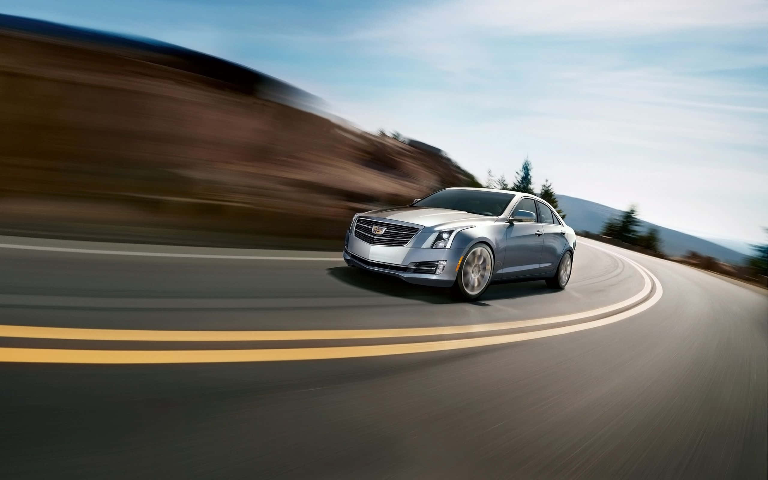 Cadillac Xts 桌布 壁紙 — 一輛銀色Cadillac轎車在彎曲的山路上疾馳，背景模糊，天空清澈湛藍。
