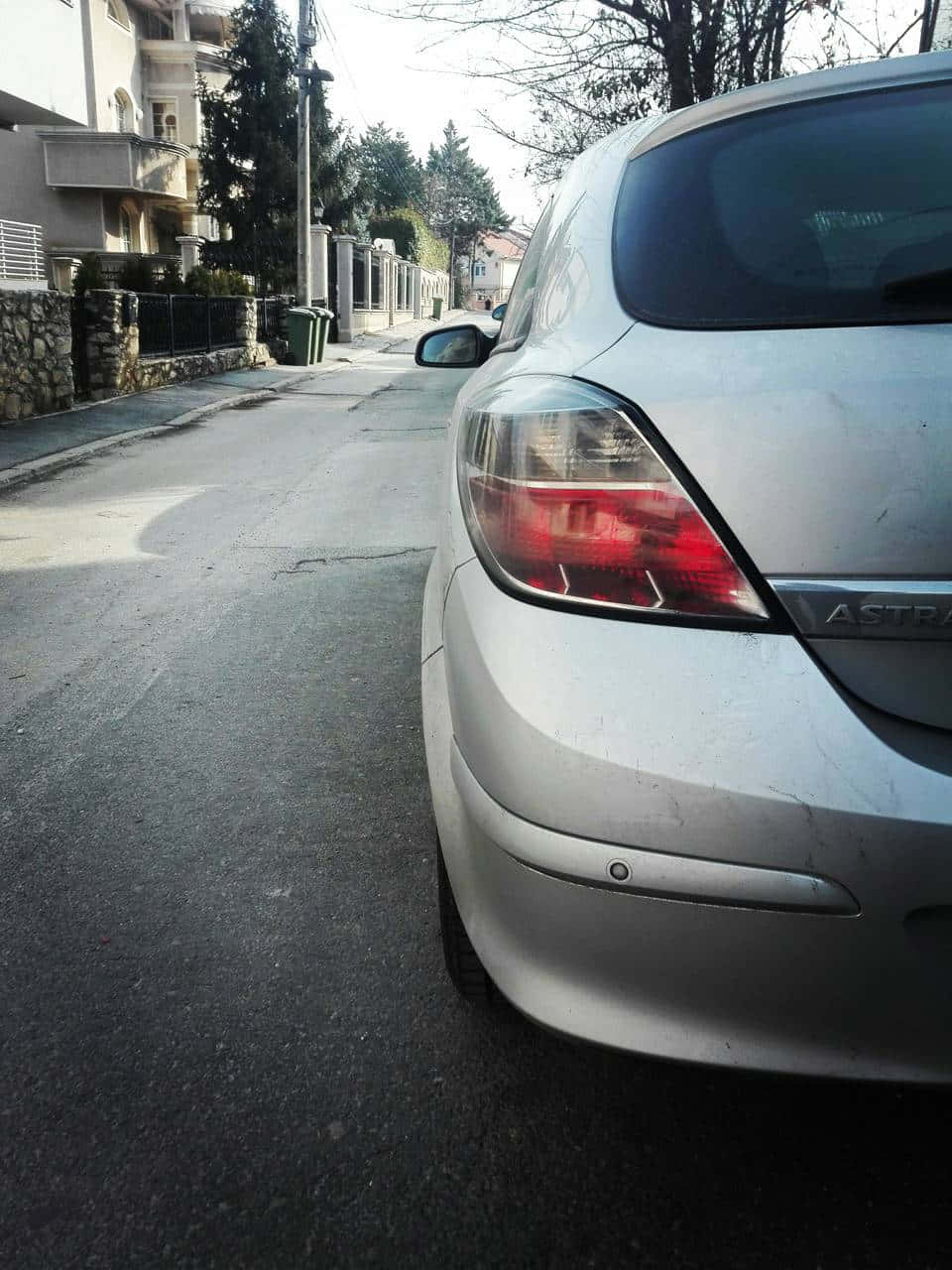 Opel Astra 桌布 壁紙 — 銀色汽車停泊在寧靜住宅區的街道上，兩旁有石牆和光禿禿的樹木，在明亮的日光下。