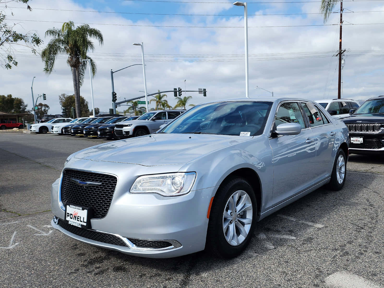 Chrysler 300 桌布 壁紙 — 一輛銀色克萊斯勒轎車在部分多雲的天空下，在繁忙的經銷商場地中閃閃發光，場地兩旁種有棕櫚樹和交通燈。