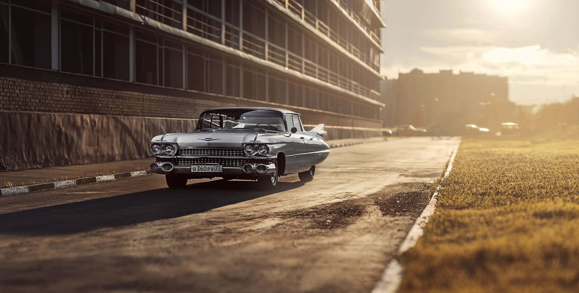 Cadillac Deville 桌布 壁紙 — 一輛銀色經典汽車在陽光明媚的城市道路上行駛，旁邊是磚砌建築和草地邊緣。
