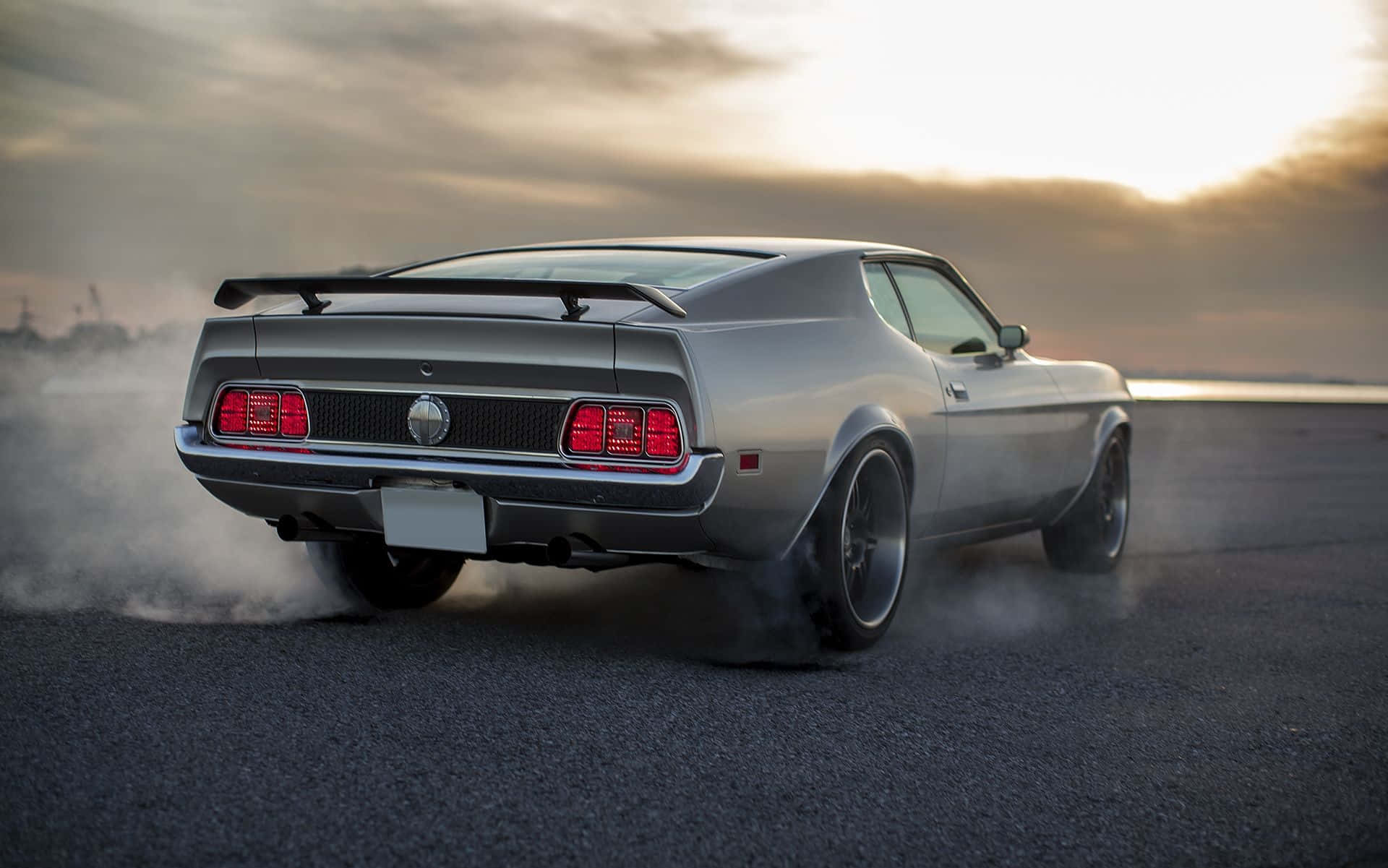 Ford Mustang Mach 1 桌布 壁紙 — 一輛銀色經典肌肉車在金色夕陽天空下漂移，後輪冒出滾滾濃煙。