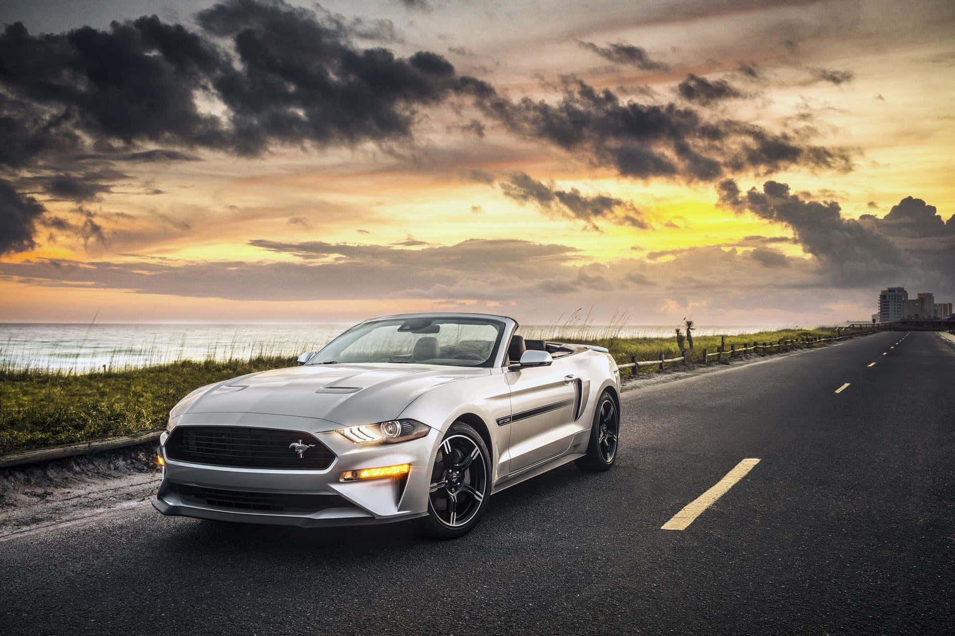 Ford Mustang California Special 桌布 壁紙 — 一輛銀色開篷跑車在日落時分沿著海岸公路行駛，背景是戲劇性的雲層和海洋。