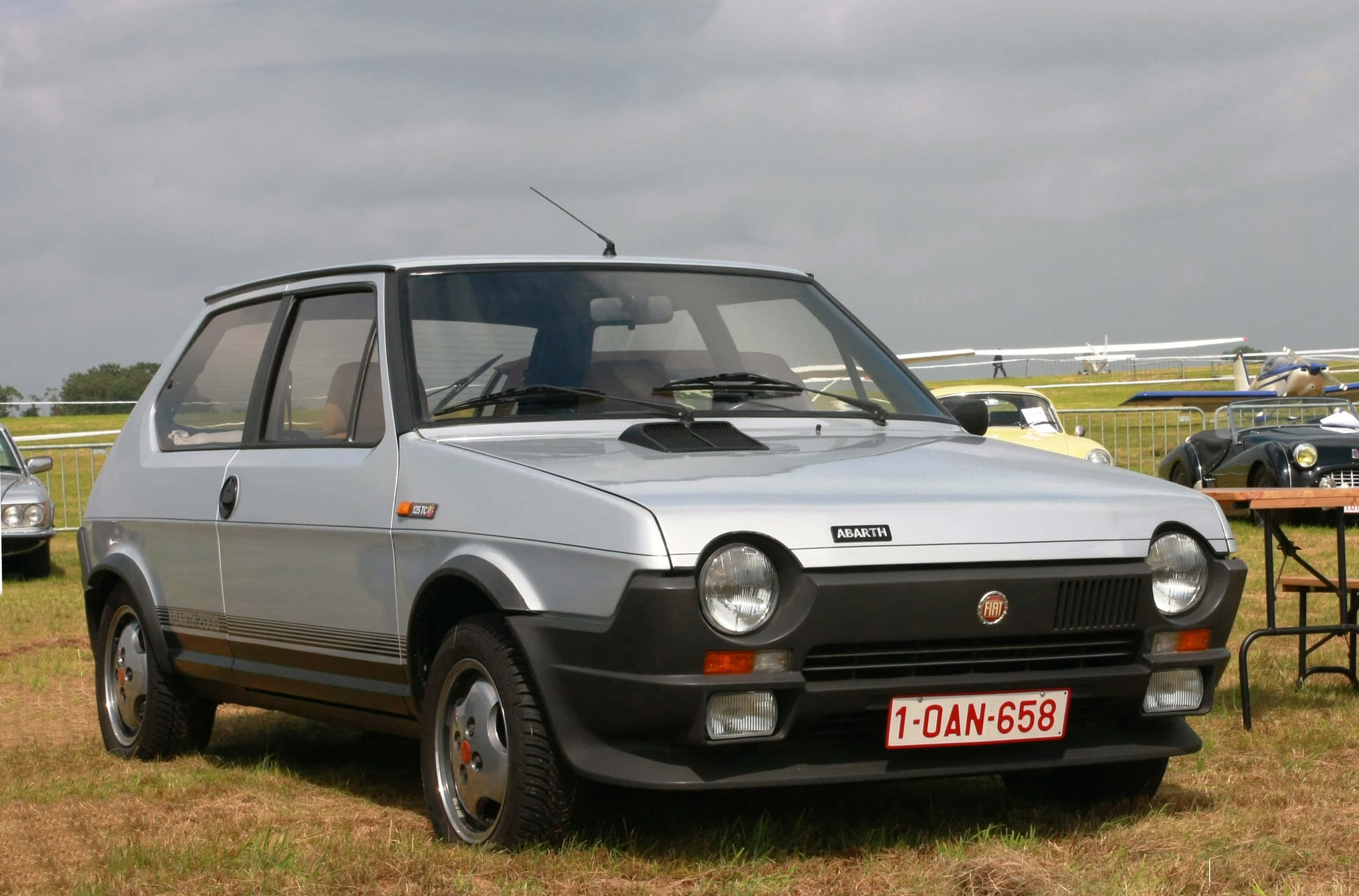 Fiat Ritmo 桌布 壁紙 — 一輛銀色嘅Fiat Abarth 124 Sport Spider停泊喺草地嘅老爺車展上，天空多雲。
