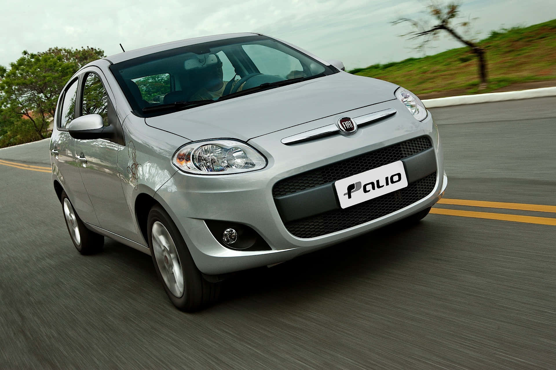 Fiat Palio 桌布 壁紙 — 一輛銀色Fiat Palio在蜿蜒的道路上疾馳，背景是模糊的綠樹和陰天。