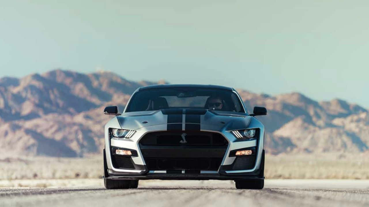  壁紙 — 一輛銀色福特野馬Shelby GT500帶有黑色賽車條紋，在晴朗天空下，背景是崎嶇山脈的沙漠道路上向前行駛。
