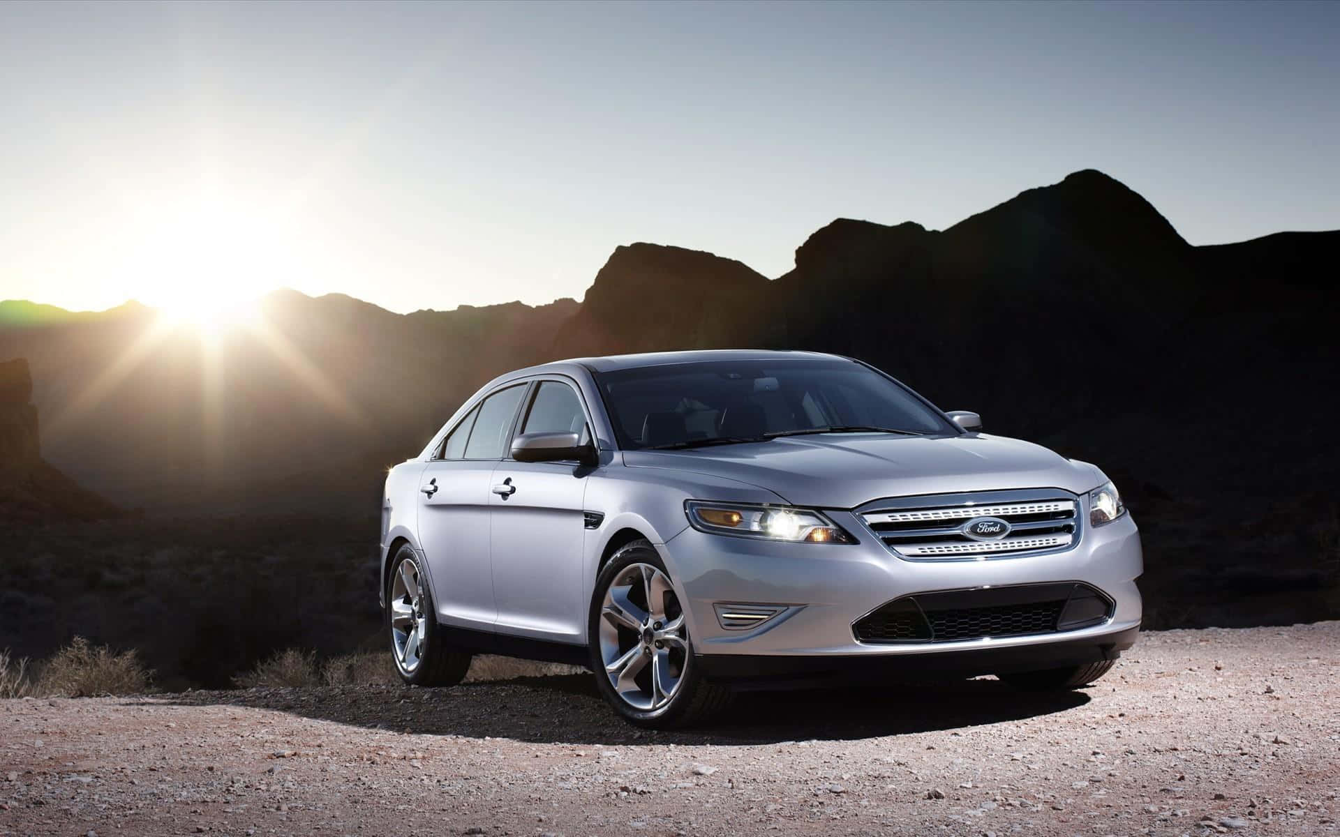 Ford Taurus 桌布 壁紙 — 一輛銀色福特Taurus轎車在明亮的陽光下，映襯著崎嶇的沙漠山脈閃閃發光。