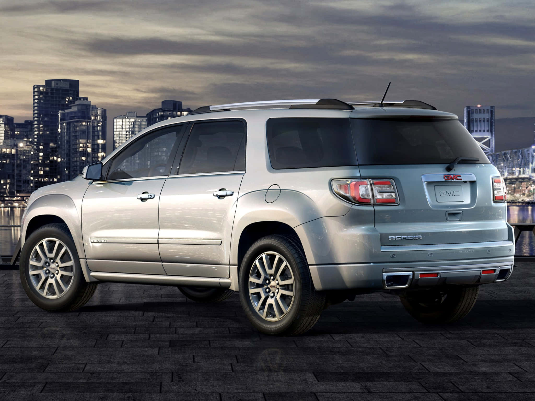 Gmc Acadia 桌布 壁紙 — 一架銀色GMC Acadia SUV停泊在深色磚砌海濱，背後的城市天際線在黃昏中閃耀。