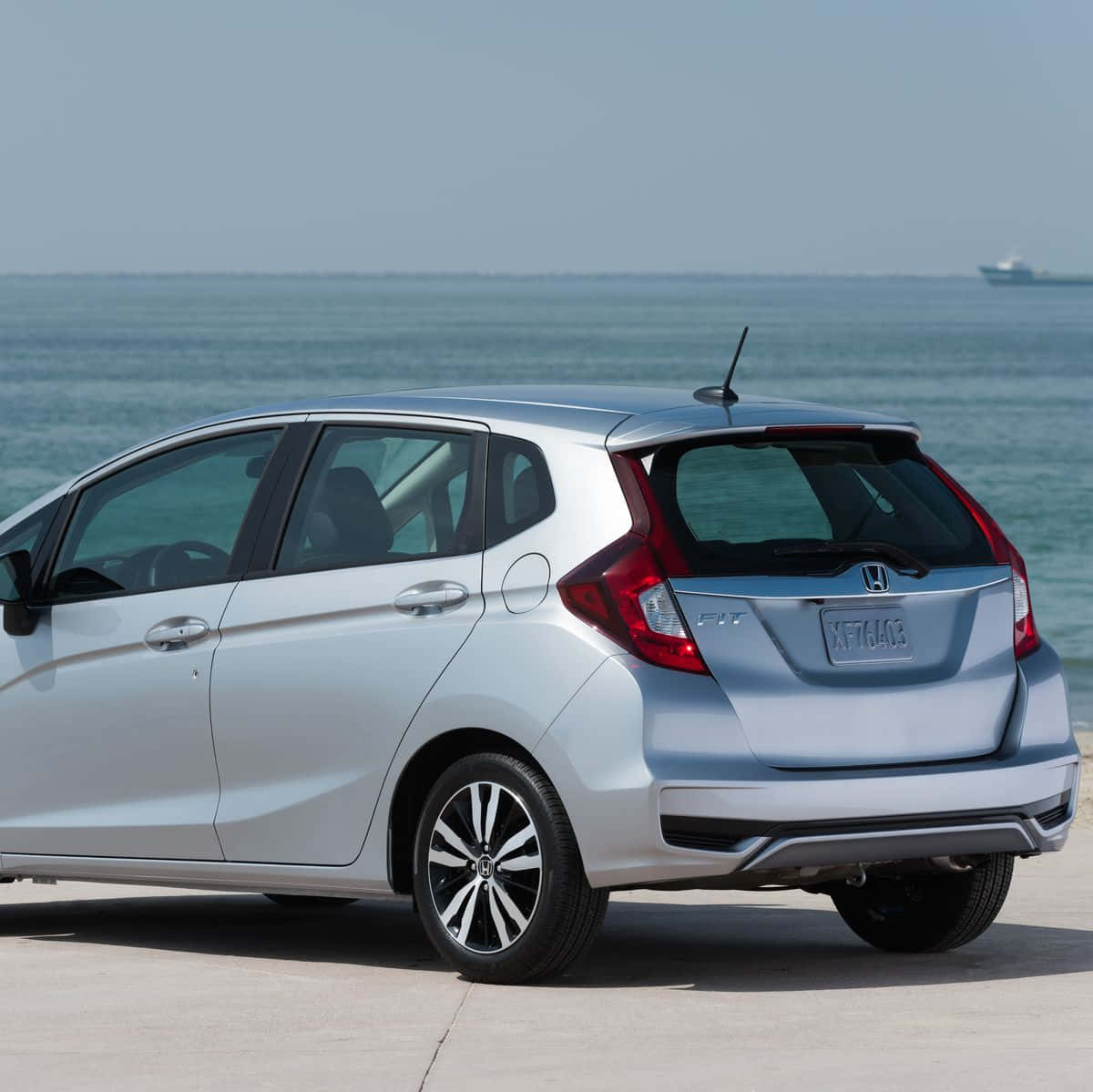 Honda Fit 桌布 壁紙 — 一輛銀色本田Fit停泊在混凝土碼頭上，海洋波浪和遠處的船隻在清澈的藍天下。