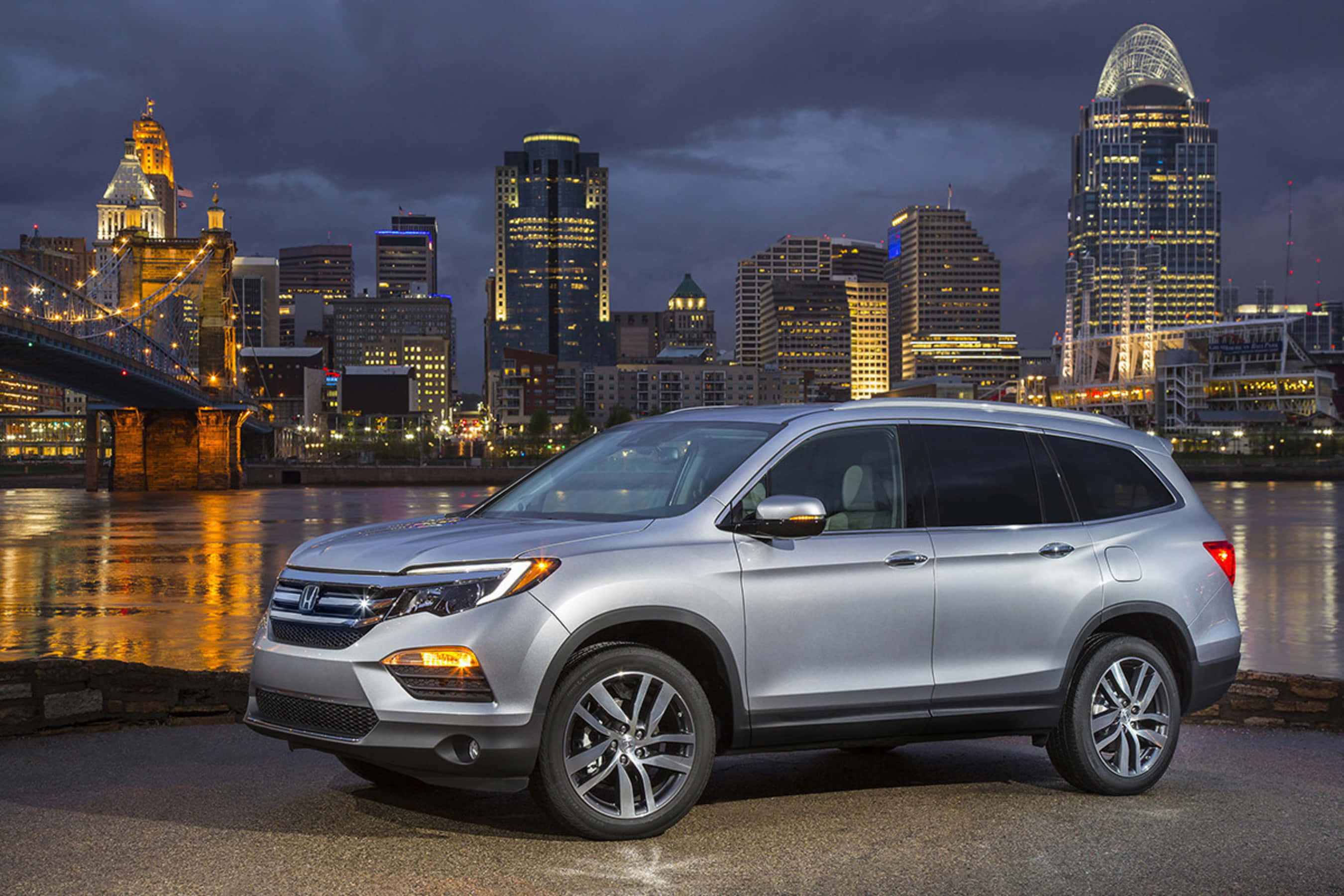 Honda Pilot 桌布 壁紙 — 一輛銀色本田Pilot SUV在黃昏時停泊在河邊，背後是充滿活力的城市天際線和發光的橋樑。