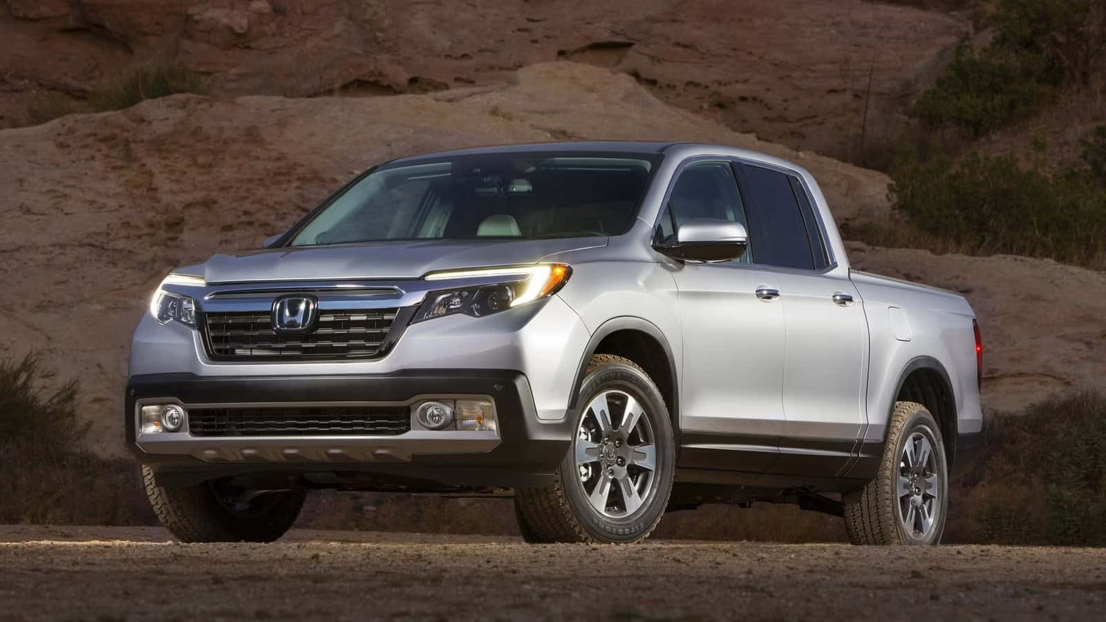 Honda Ridgeline 桌布 壁紙 — 一架銀色本田 Ridgeline 貨卡停泊在塵土飛揚的沙漠道路上，背景是岩石地形和稀疏的綠色植物。