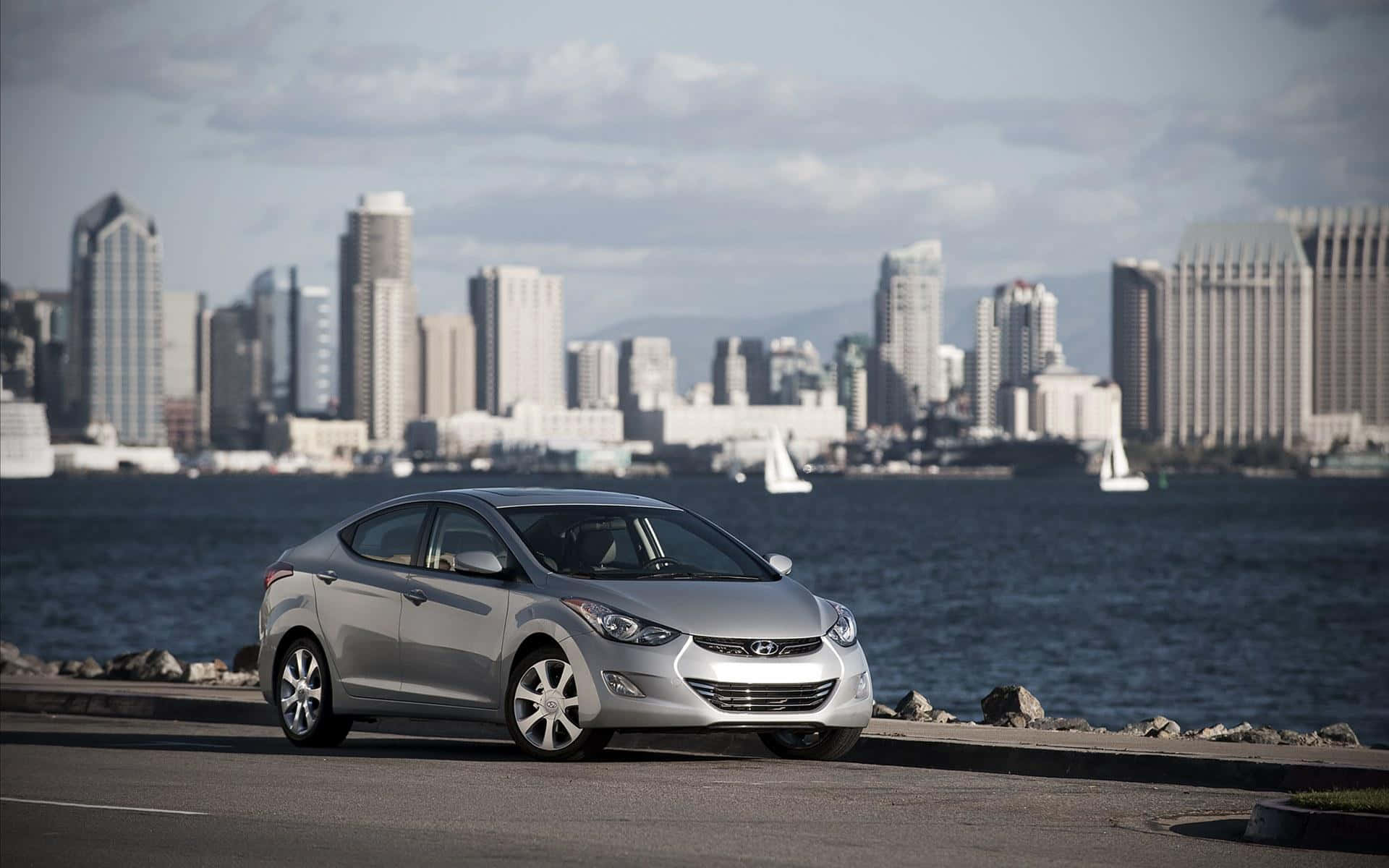Hyundai Elantra 桌布 壁紙 — 一輛銀色現代伊蘭特停在海濱，背景是城市天際線和帆船。