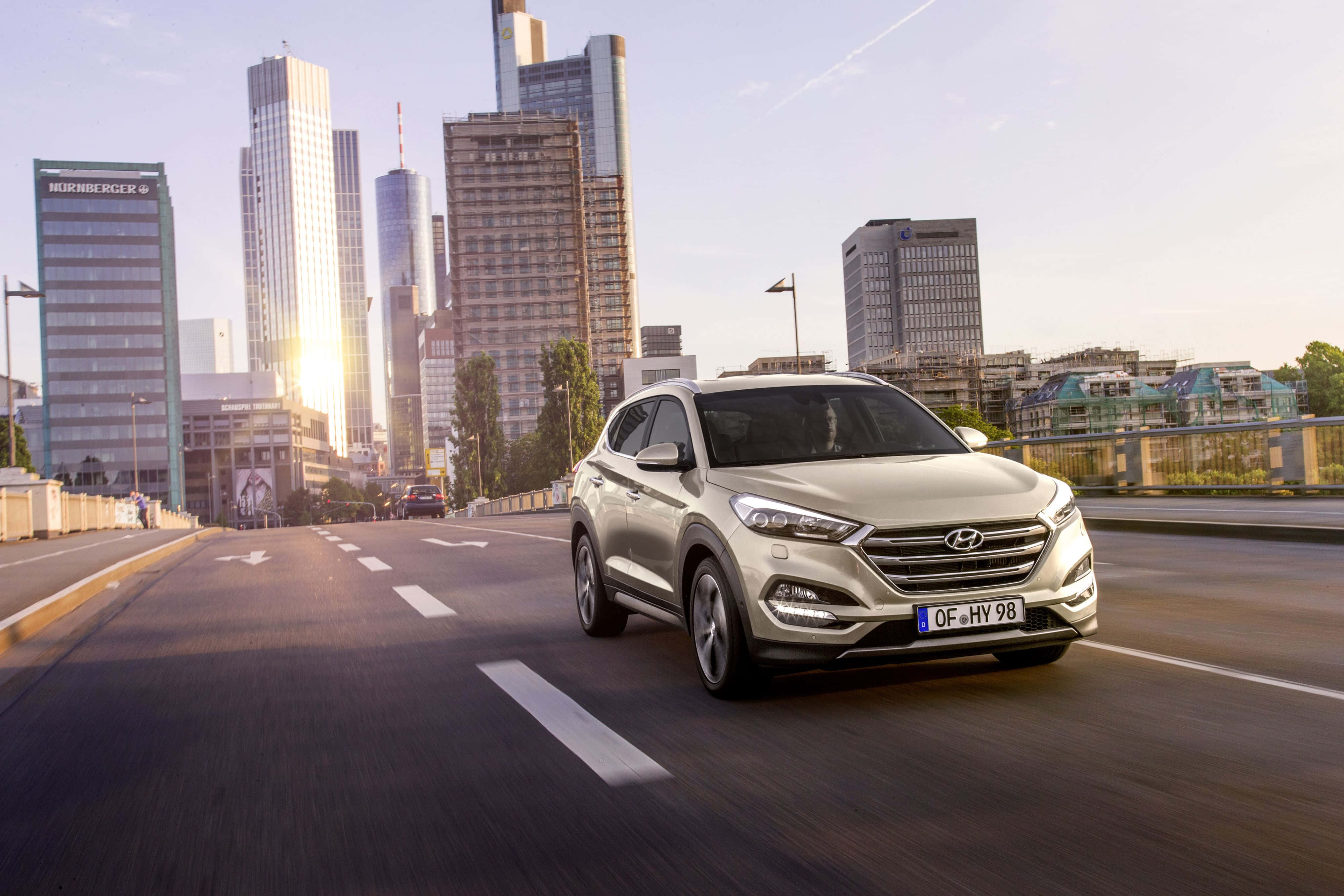 Hyundai Tucson 桌布 壁紙 — 一輛銀色現代SUV在城市的橋上行駛，現代摩天大樓在金色的日落光芒中閃耀。
