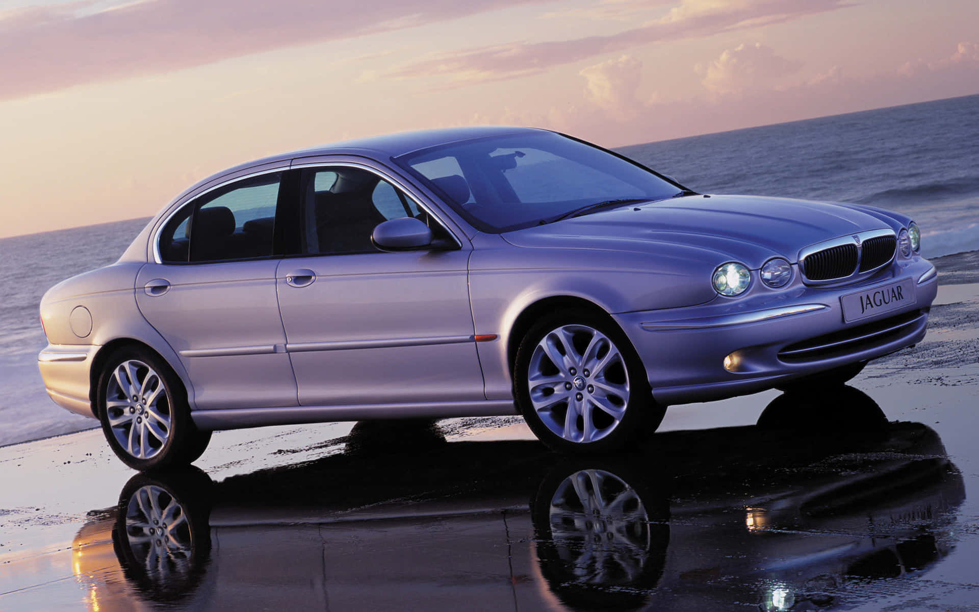 Jaguar X-type 桌布 壁紙 — 一輛銀色積架房車在日落時分在濕沙上閃閃發光，映照著海浪和天空。