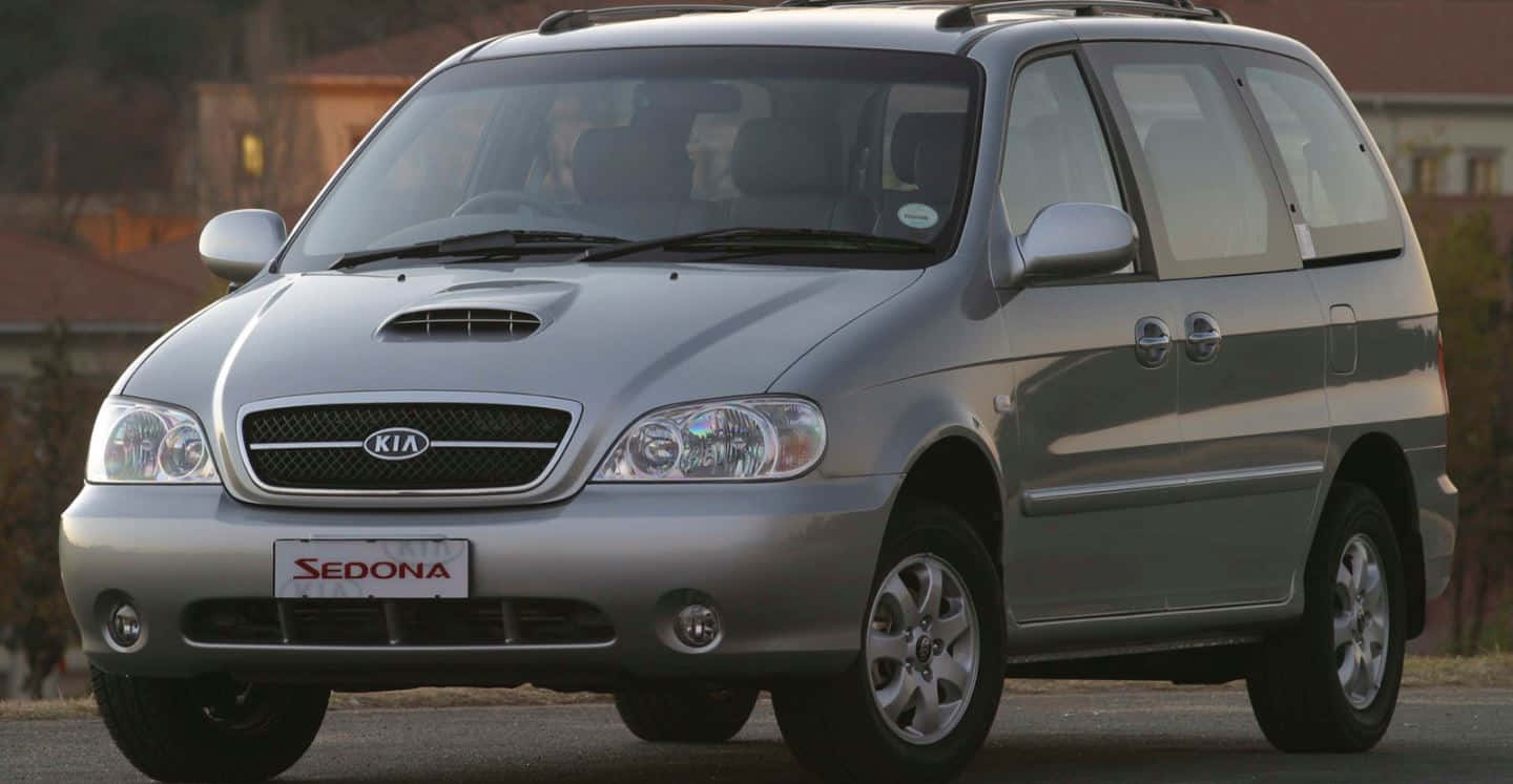 Kia Sedona 桌布 壁紙 — 一輛銀色Kia Sedona小型貨車停泊在街道上，背景有房屋，在柔和日光下。
