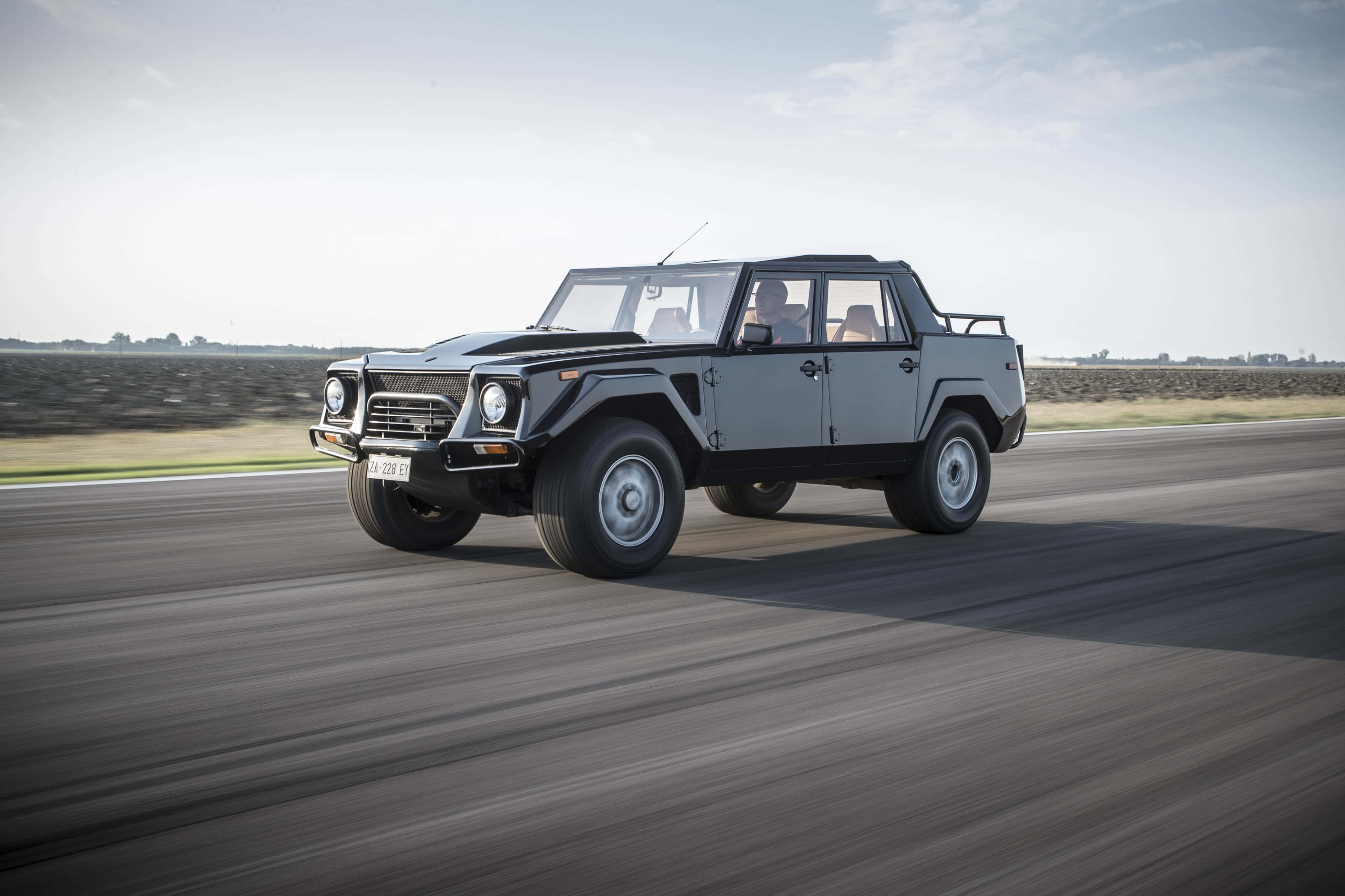 Lamborghini Lm002 桌布 壁紙 — 一輛銀色林寶堅尼LM002 SUV在陽光照耀的瀝青路上飛馳，背景是模糊的動態和開闊的田野。