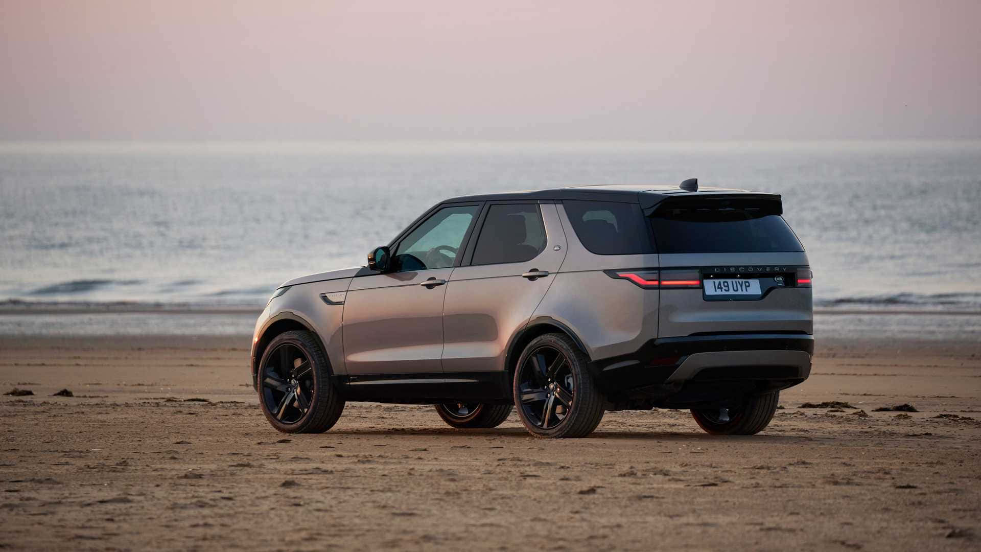 Land Rover Discovery 桌布 壁紙 — 一輛時尚的銀色 Land Rover Discovery 停泊在沙灘上，背景是平靜的海浪和日落時柔和的粉紅色天空。