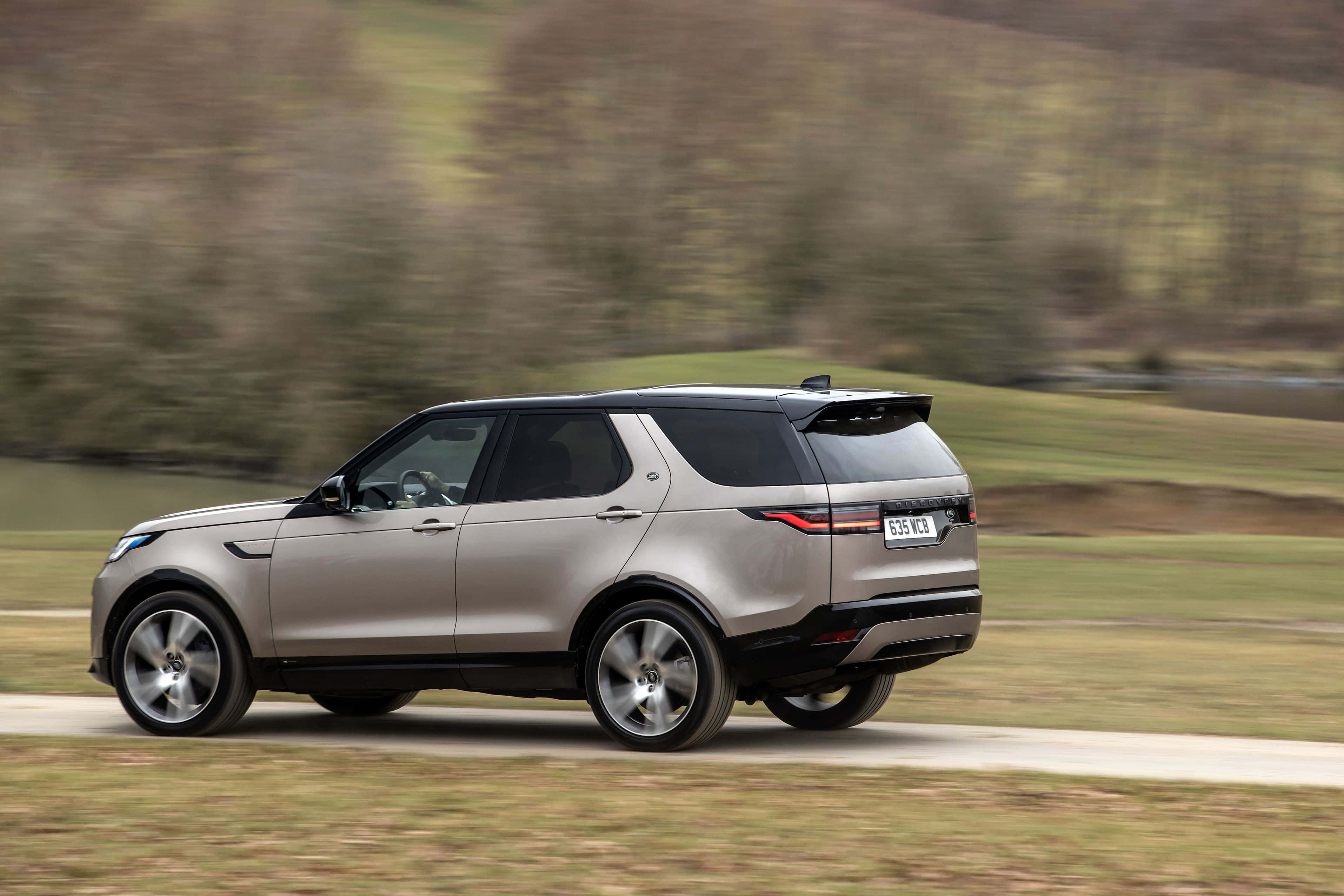 Land Rover Discovery 桌布 壁紙 — 一輛銀色 Land Rover Discovery SUV 在鄉村道路上飛馳，背景是模糊的綠色山丘。