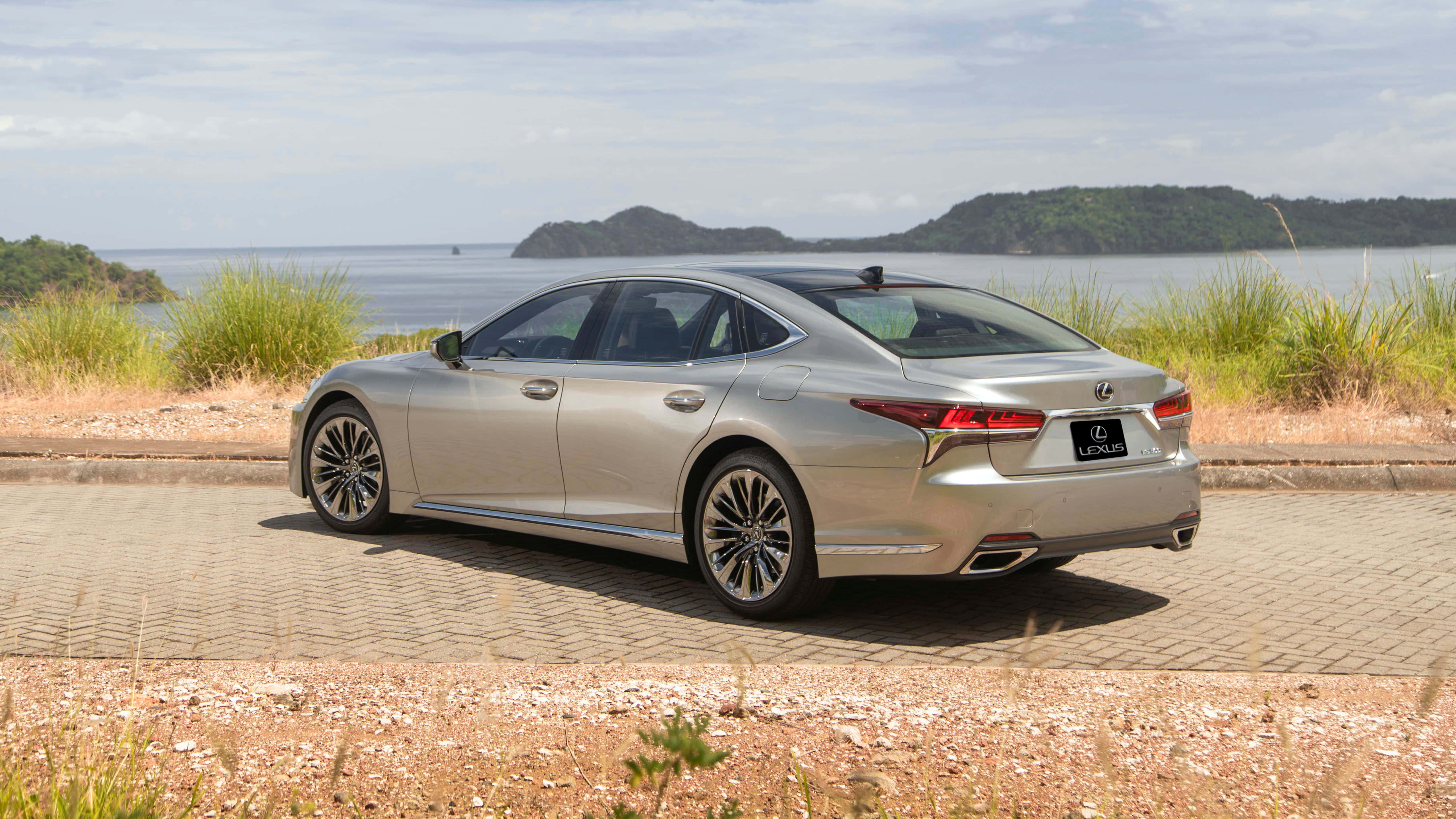 Lexus Ls 500 桌布 壁紙 — 一架流線型銀色Lexus房車停泊在鋪砌道路上，旁邊是草叢生的岸邊，遠處有島嶼，天空多雲。