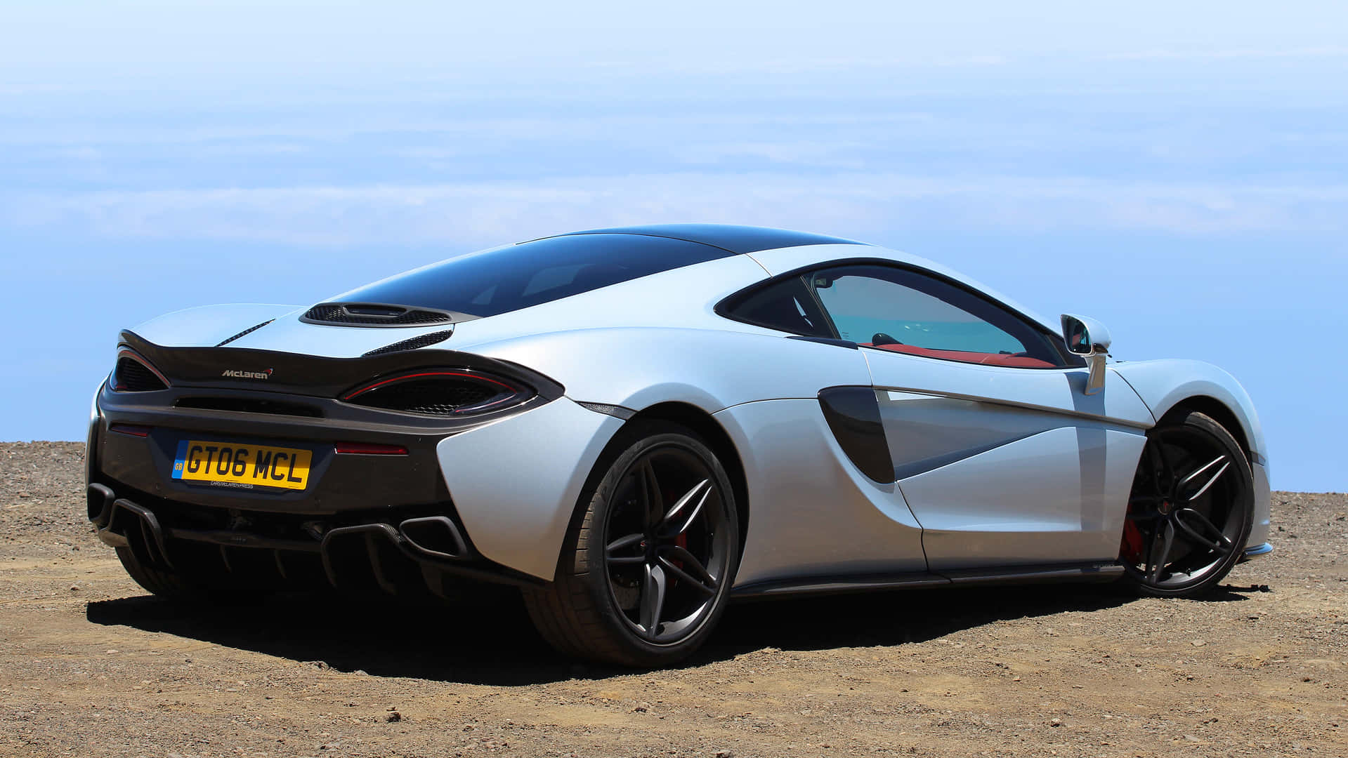 Mclaren 570gt 桌布 壁紙 — 一架流線型銀色McLaren跑車停泊在俯瞰海洋的塵土懸崖上，天空清澈湛藍。
