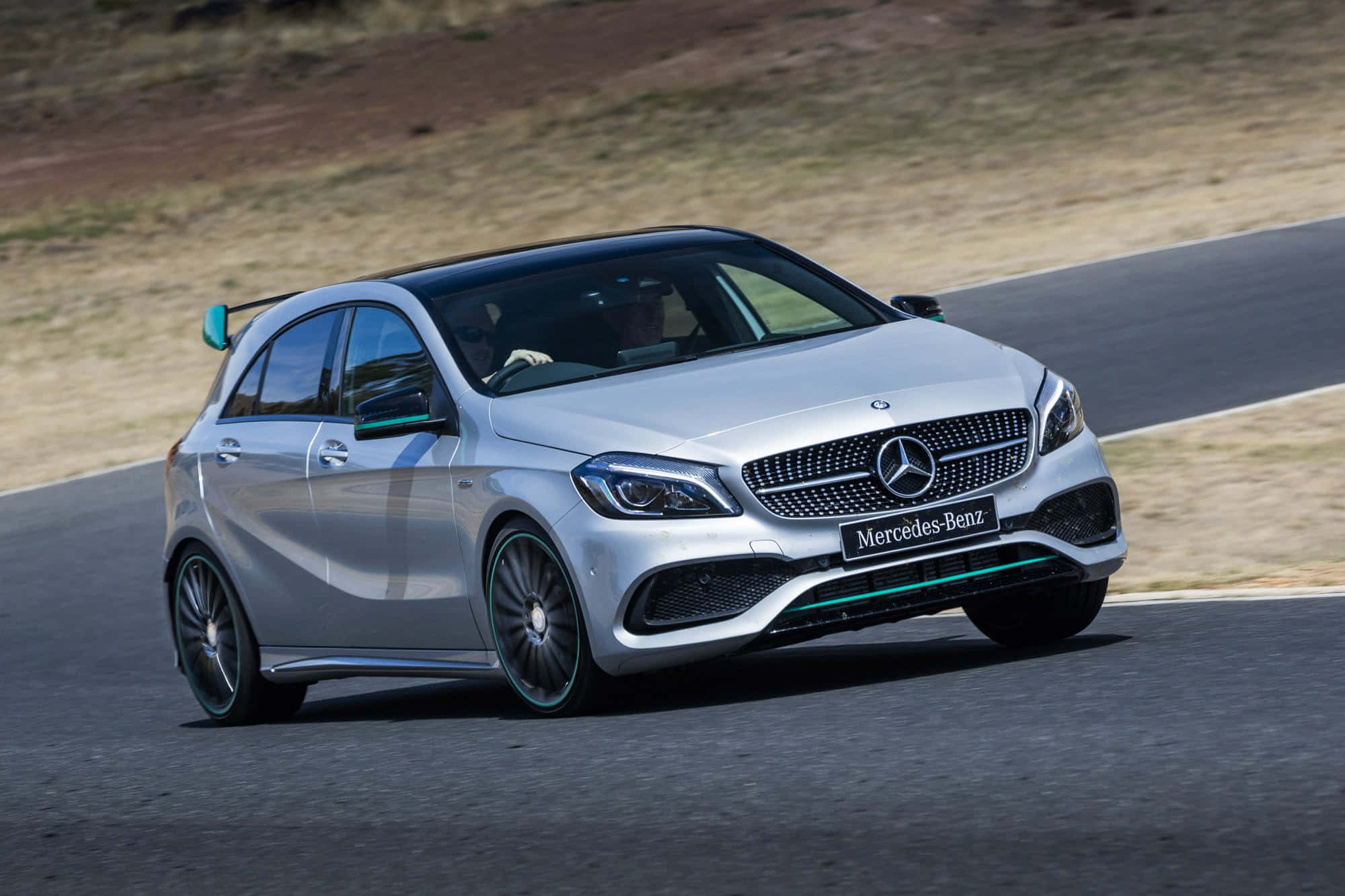 Mercedes Benz A-class 桌布 壁紙 — 一輛銀色 Mercedes-Benz A45 AMG 在蜿蜒的瀝青路上疾馳，背景是乾燥的草坡。