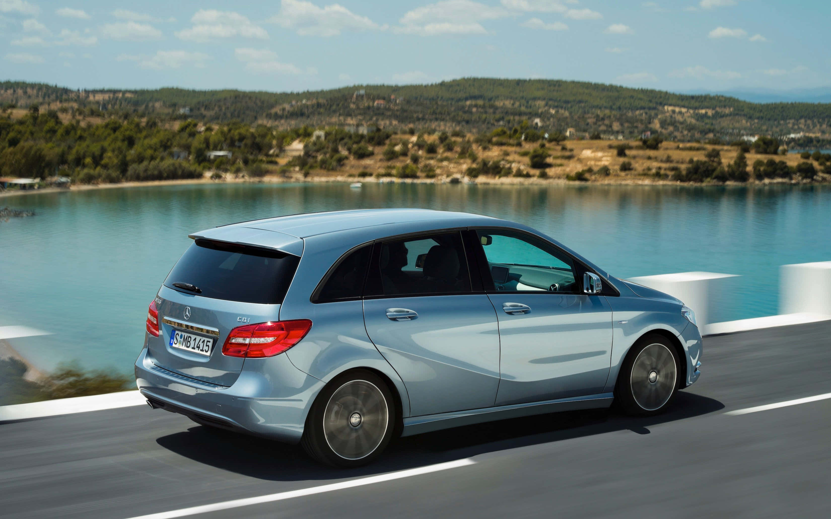 Mercedes Benz B-class 桌布 壁紙 — 一輛時尚的銀色 Mercedes-Benz B-Class 沿著海岸道路行駛，旁邊是碧綠的海水，頭上是明亮的藍天。