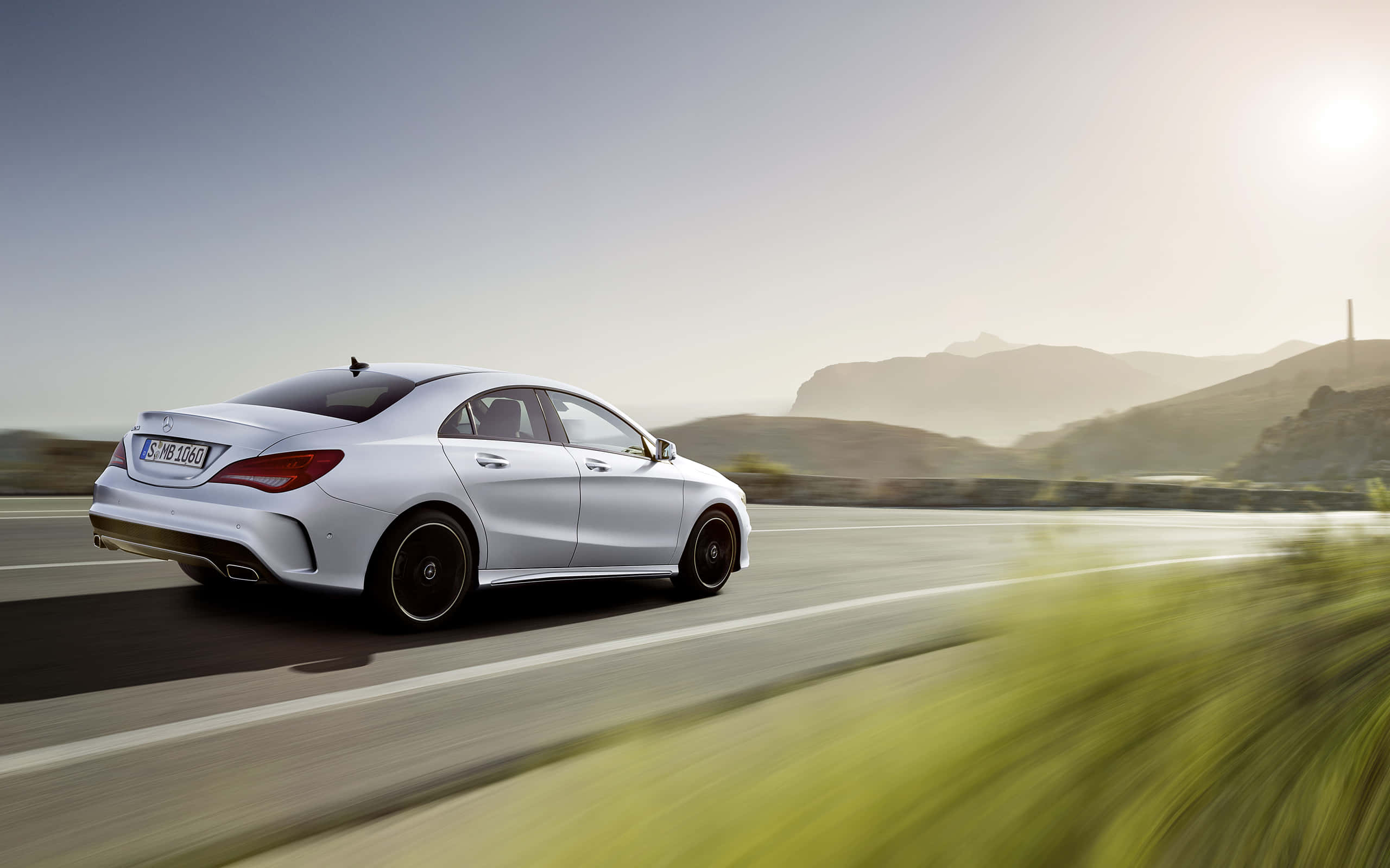Mercedes Benz Cla-class 桌布 壁紙 — 一輛時尚的銀色 Mercedes-Benz CLA 轎車在陽光普照的山丘間沿著蜿蜒的道路疾馳，周圍綠意朦朧。