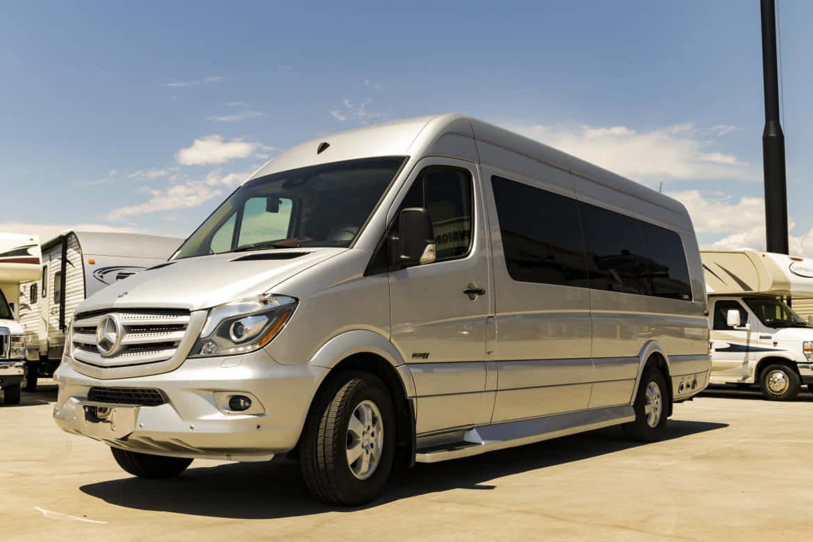 Mercedes Benz Sprinter 桌布 壁紙 — 一架時尚的銀色 Mercedes-Benz Sprinter 客貨車停泊在陽光明媚的停車場，背景有露營車，天空蔚藍。