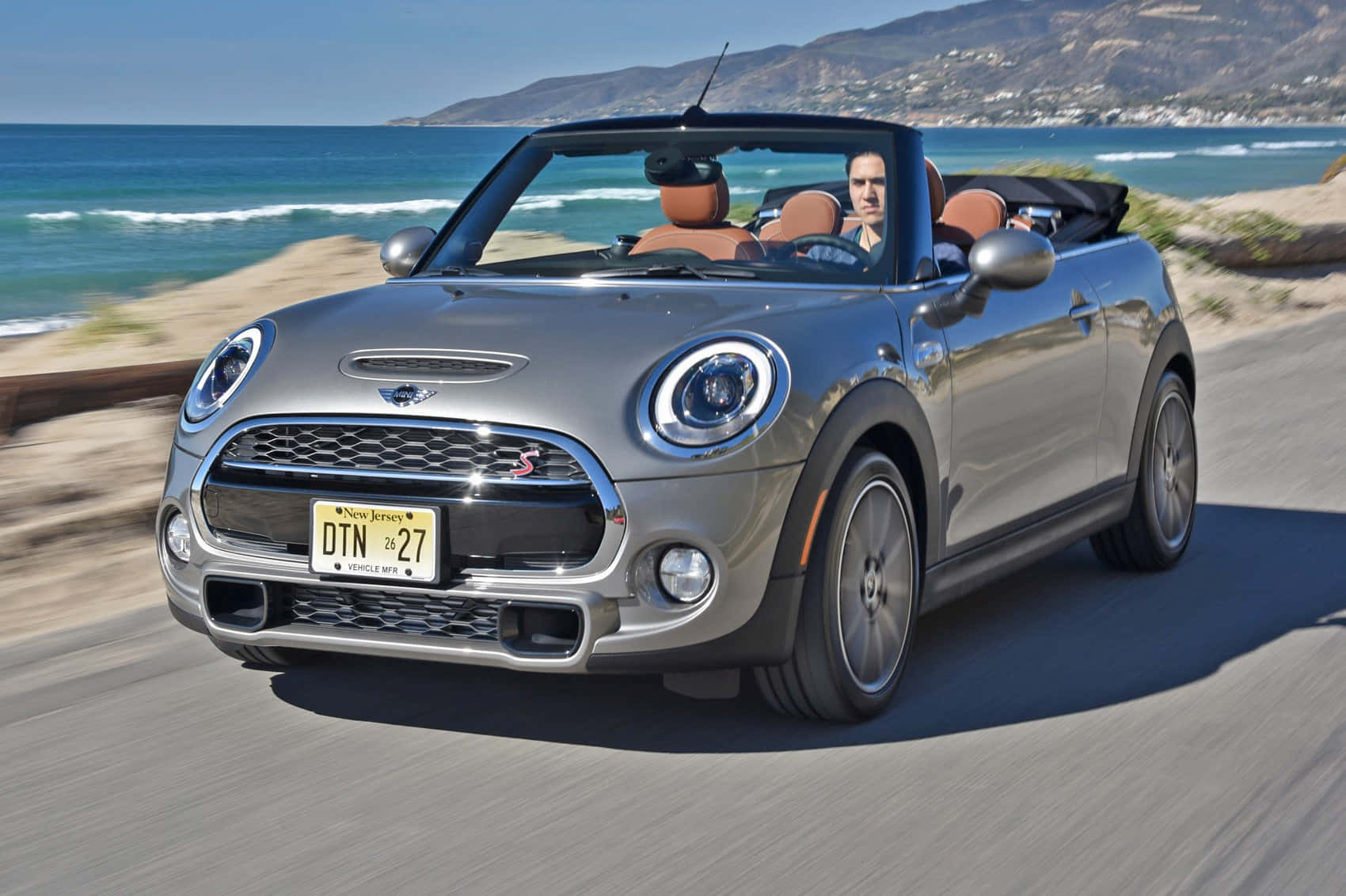 Mini Cooper Convertible 桌布 壁紙 — 一輛銀色MINI Cooper開篷跑車沿海岸公路飛馳，背景有海浪和山脈。