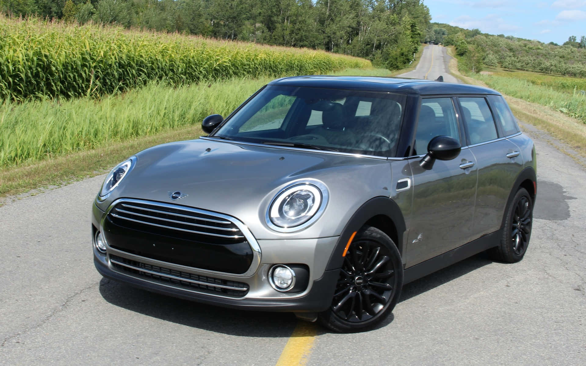 Mini Cooper S Clubman All4 桌布 壁紙 — 一輛時尚的銀色MINI Countryman在晴朗藍天下，行駛在兩旁高聳綠色玉米田的鄉村道路上。