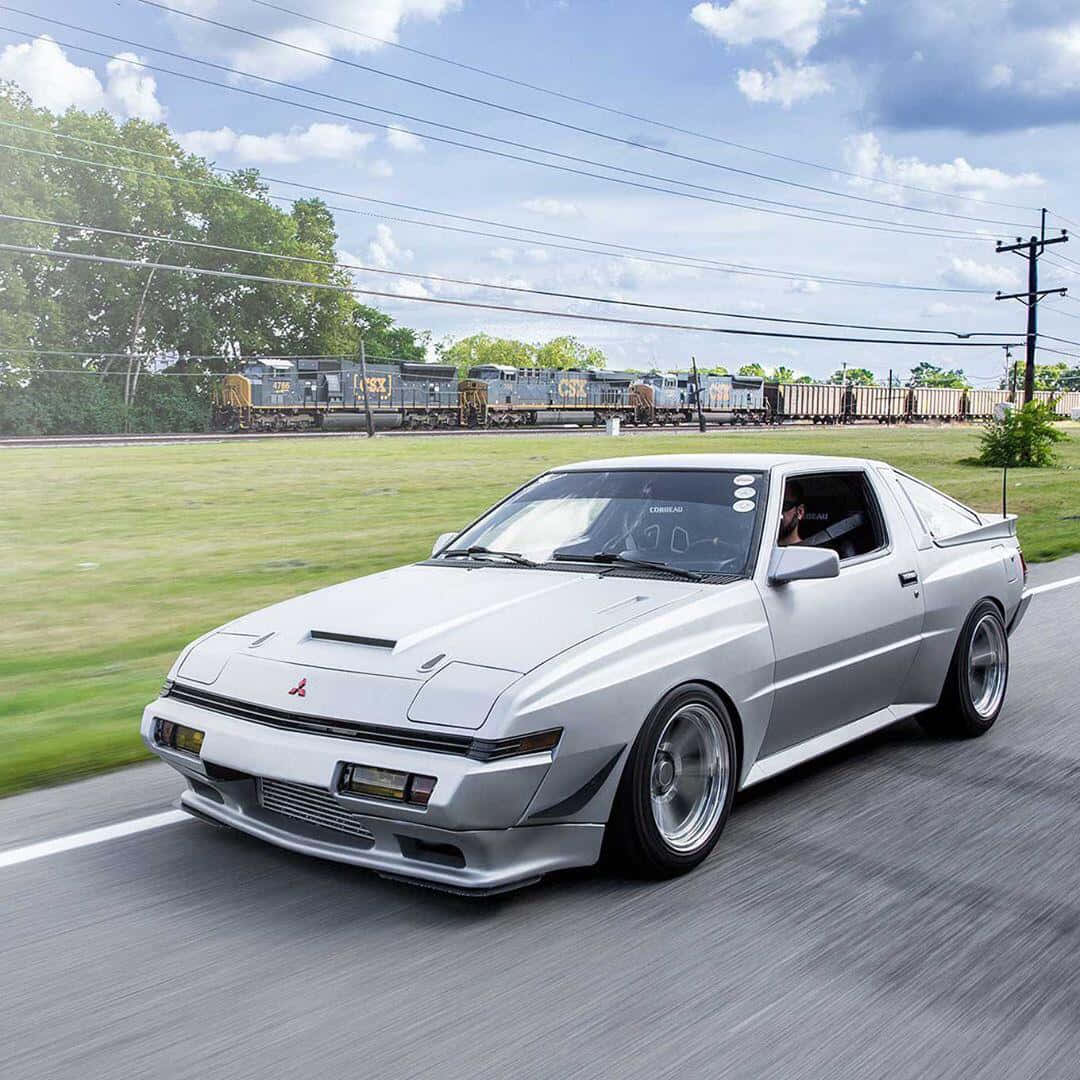 Mitsubishi Starion 桌布 壁紙 — 一輛銀色三菱Starion沿路邊飄移，背景中一列貨運火車在部分多雲的天空下駛過。