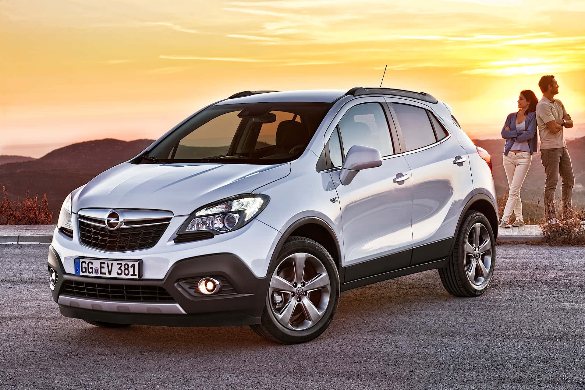 Opel Mokka 桌布 壁紙 — 一輛銀色Opel Mokka SUV停泊在日落時分的碎石路上，一對情侶站在附近觀賞金色的天空。
