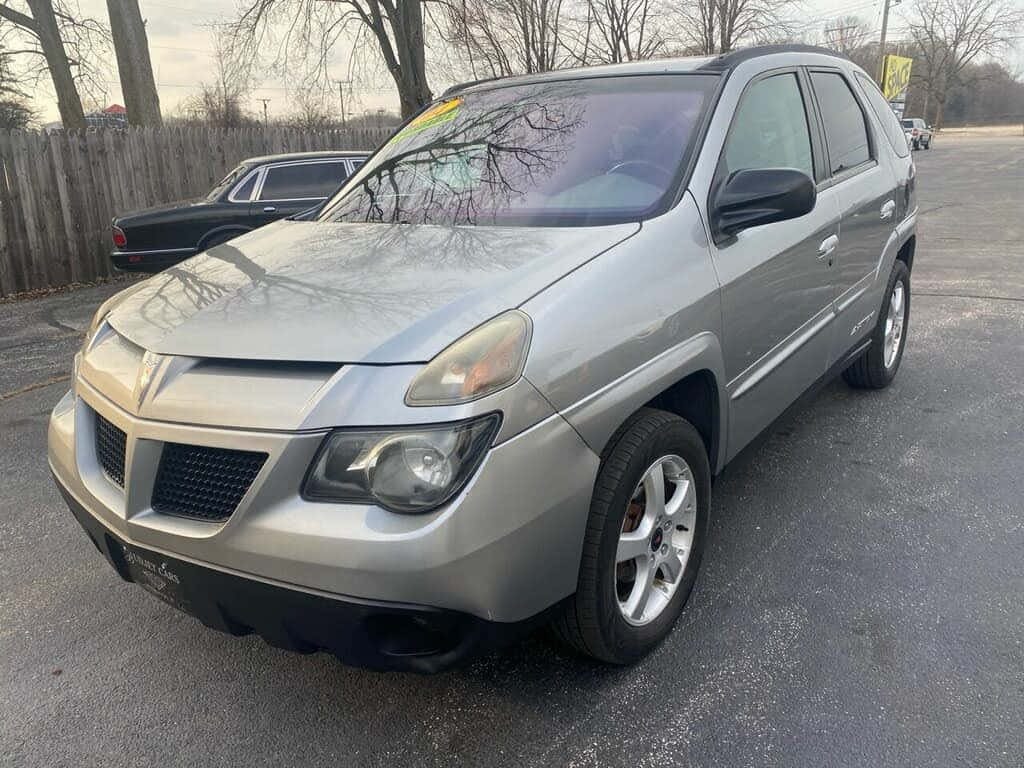  壁紙 — 一輛銀色 Pontiac Aztek SUV 停泊在瀝青路上，背景有光禿禿的樹木和木柵欄，天空多雲。