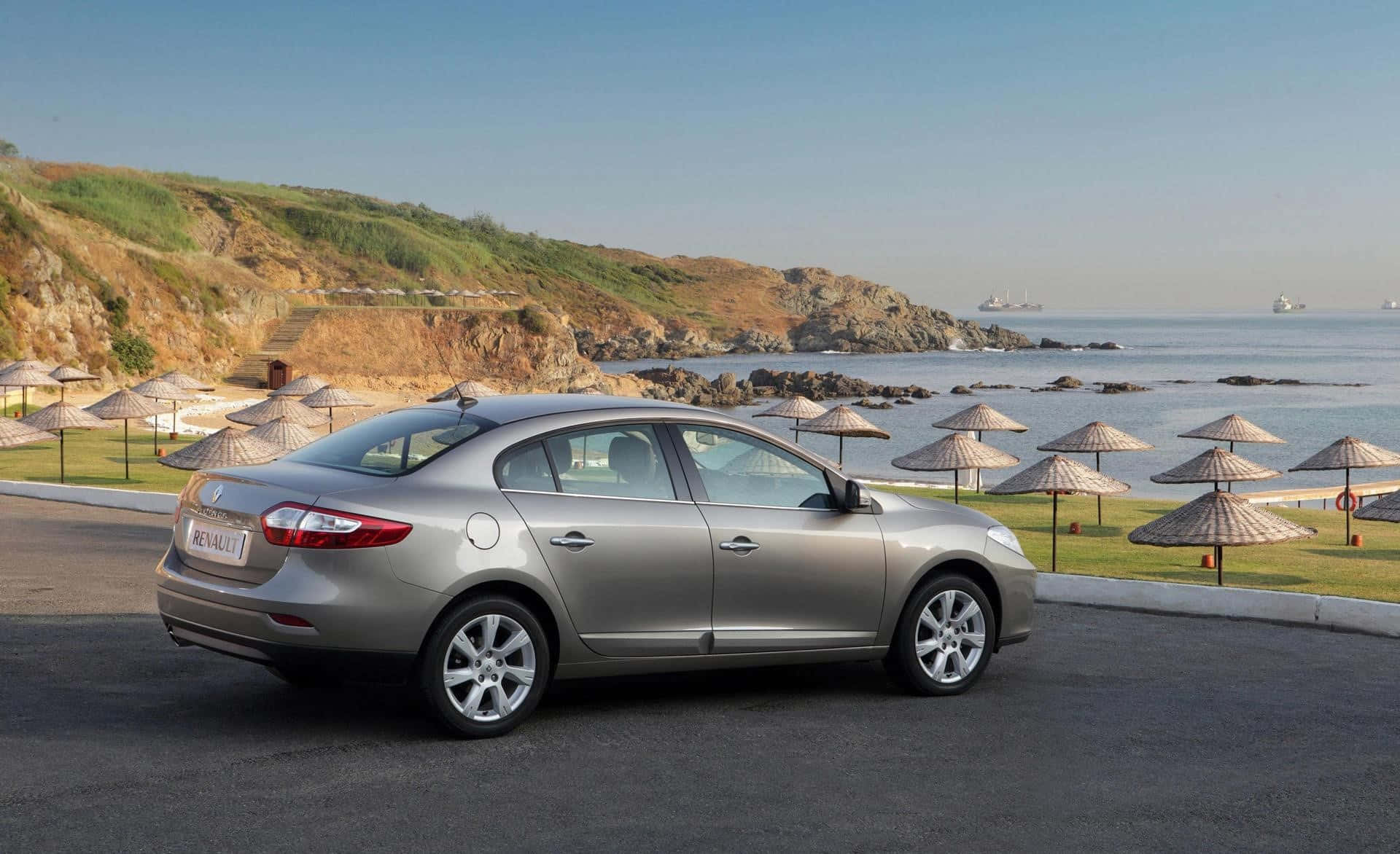 Renault Fluence 桌布 壁紙 — 一輛銀色轎車停泊在沿海公路上，旁邊是沙灘，沙灘上有茅草傘和岩石峭壁，天空清澈湛藍。