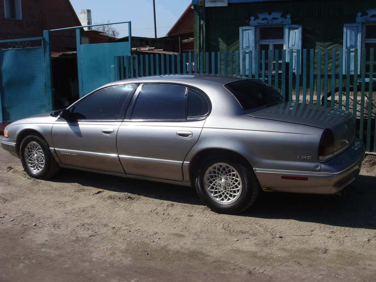 Chrysler Lhs 桌布 壁紙 — 一輛銀色房車停泊在泥地上，旁邊是青綠色的圍欄，背景有質樸的建築物，在明亮的日光下。