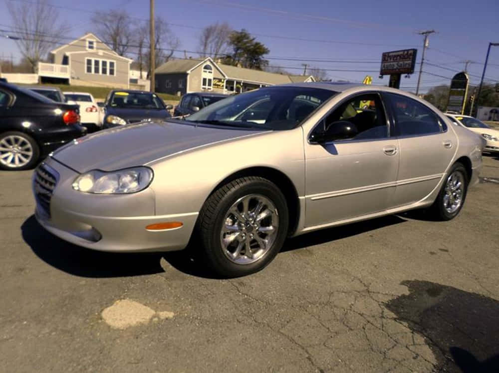 Chrysler Lhs 桌布 壁紙 — 一輛銀色房車停泊在陽光普照的二手車場，地面有裂開的瀝青，背景是住宅房屋。
