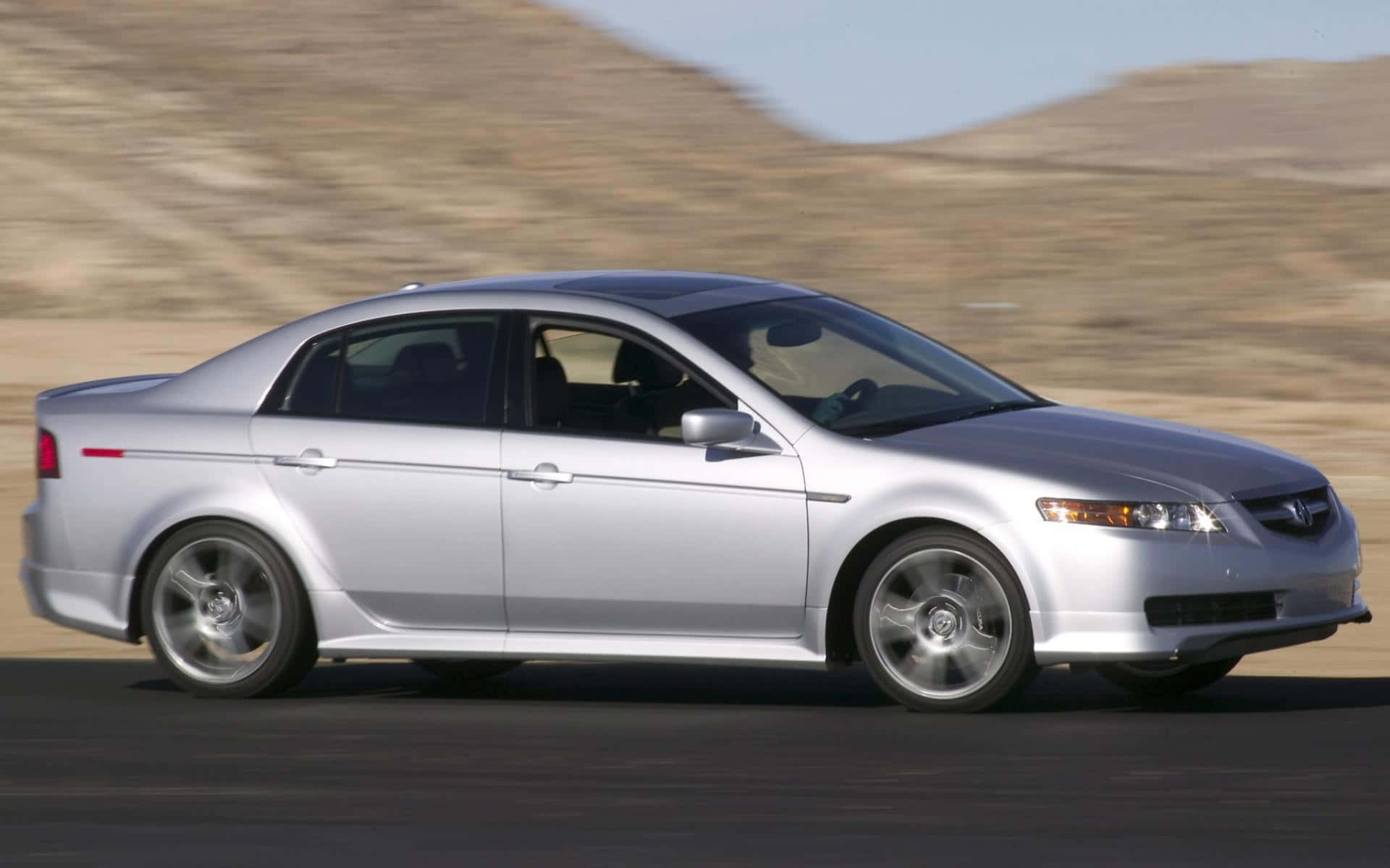Acura Tl 桌布 壁紙 — 一輛銀色轎車在沙漠公路上飛馳，背景是模糊的山丘和晴朗的天空。